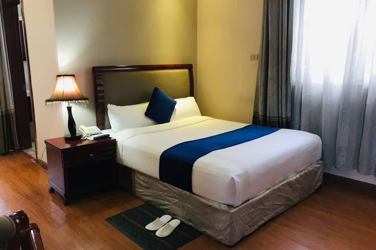 Una cama bien hecha con una almohada azul decorativa, una lámpara de noche y un par de pantuflas sobre una alfombra.
