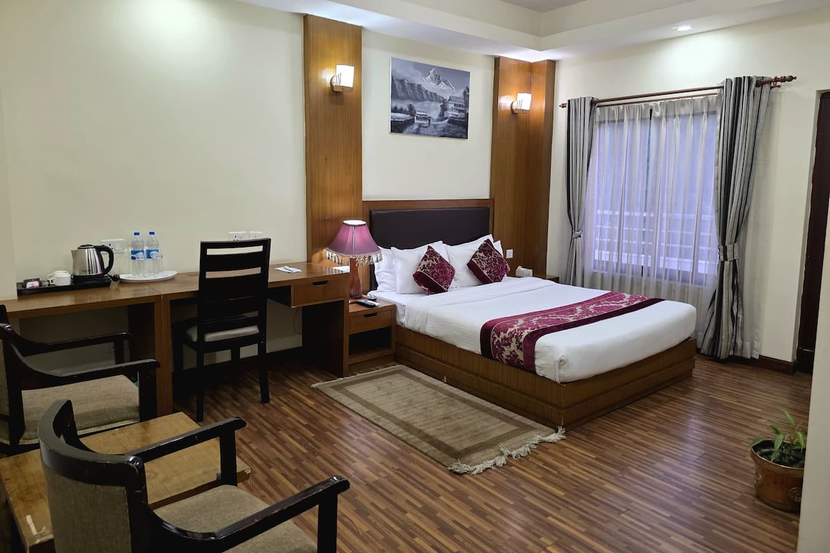 A hotel room with a wooden floor, a bed with white and maroon bedding, a desk with a lamp, a chair, and a small potted plant.