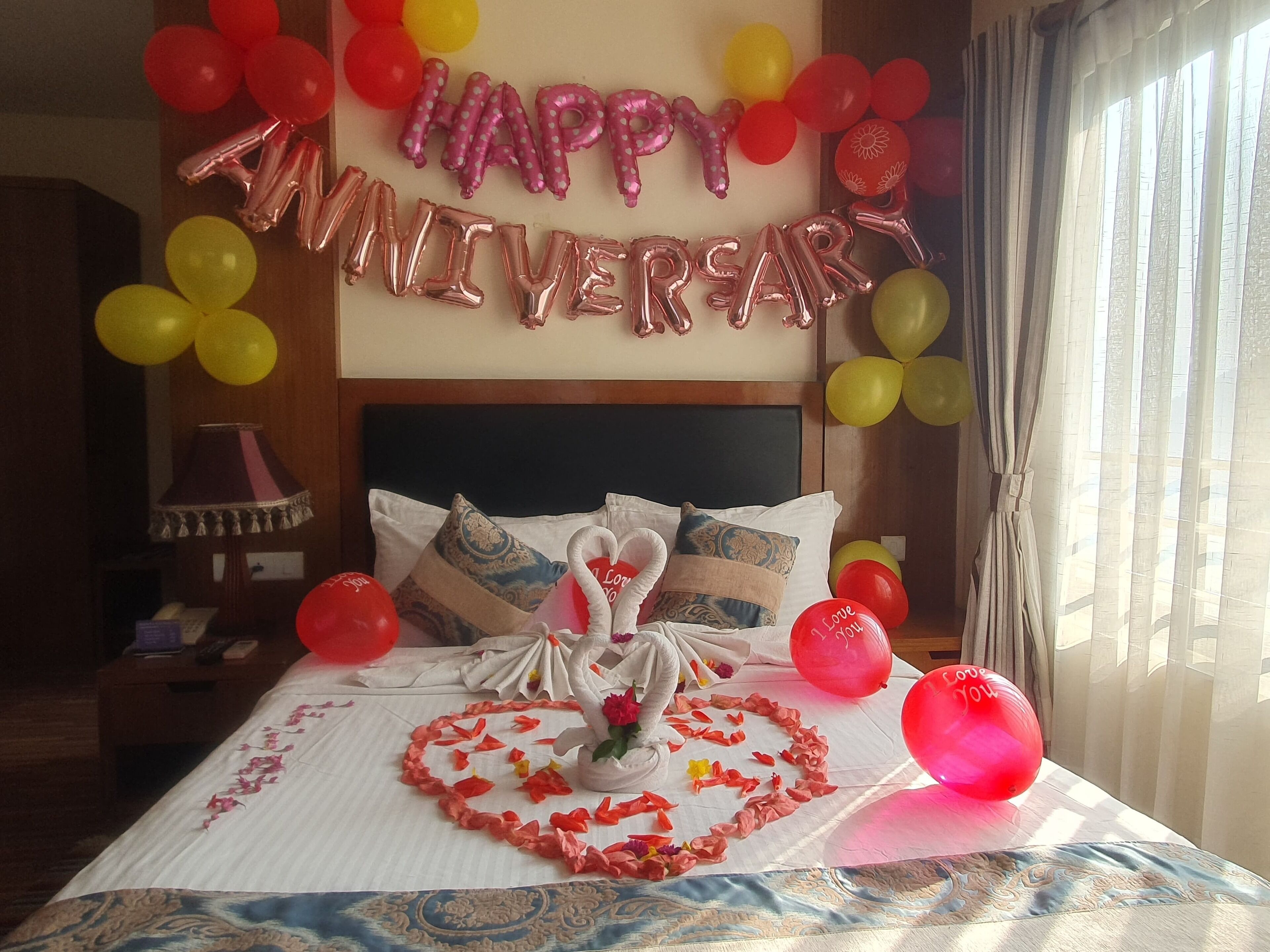 Ein Hotelzimmer mit einem Bett, das mit einer Schwanverzierung, herzförmigen Blütenblättern und Ballons geschmückt ist. Über dem Bett ist ein Luftballon-Banner mit der Aufschrift 'Happy Anniversary' sichtbar.