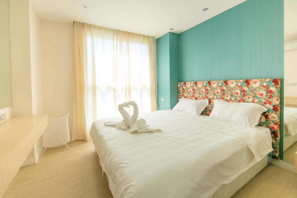 Une chambre d’hôtel avec un grand lit, une tête de lit à motifs floraux et une vue sur l’extérieur à travers des rideaux transparents.