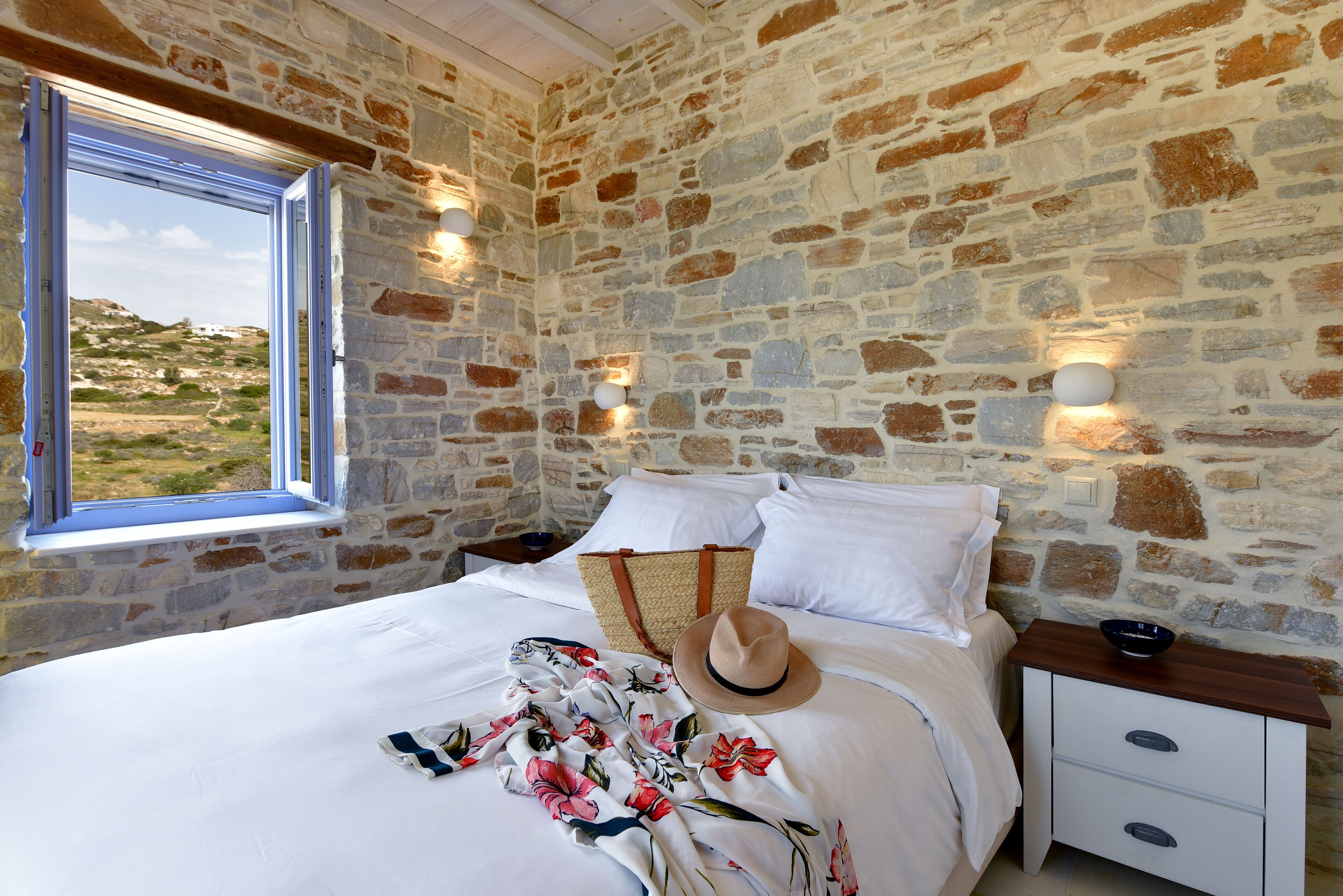 Ein Schlafzimmer mit einer Steinwand, einem Bett mit weißen Leinentüchern, einer floralen Decke, einem Hut und einem Nachttisch mit einer Schale.