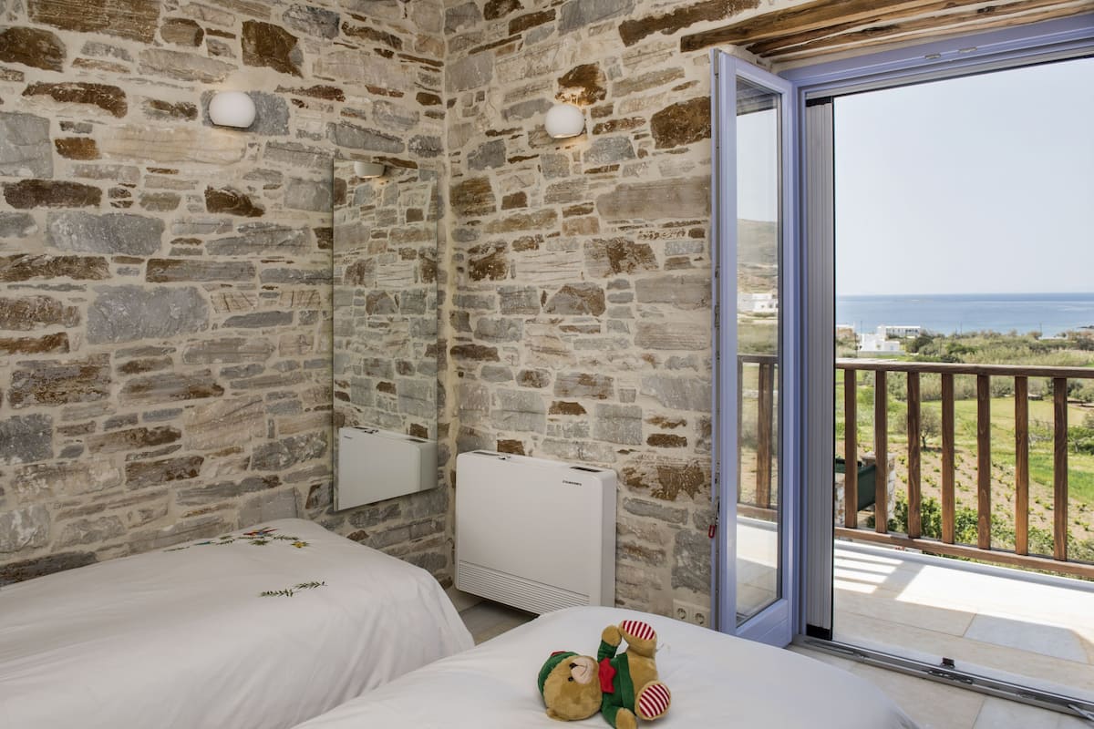 A room with stone walls, a bed, a window with a view of the sea, and a teddy bear on the bed.