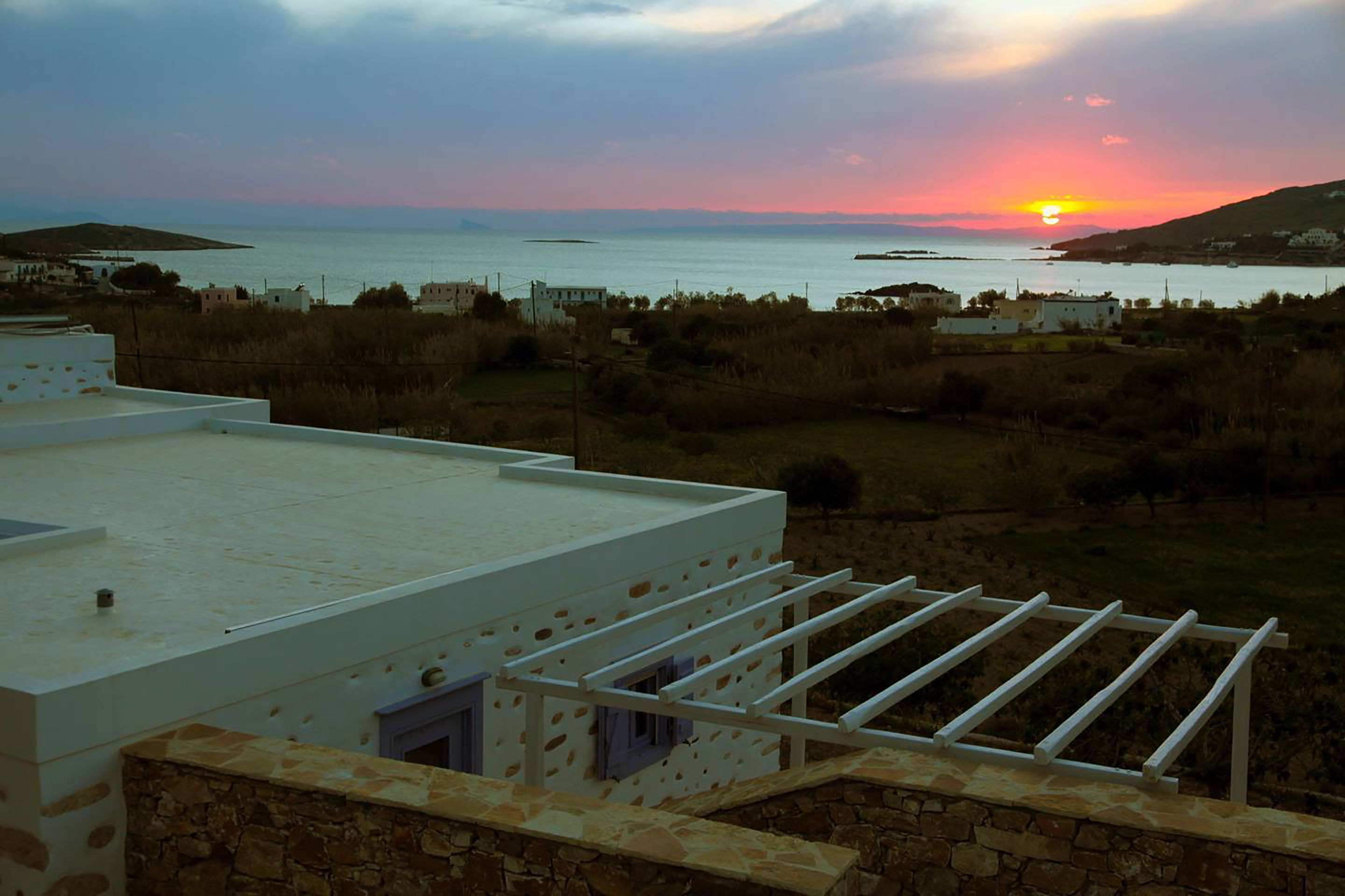 A coastal view with a sunset over the sea, a white building with a stone wall, and a white balcony with a drying rack.