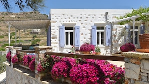 Un edificio blanco con contraventanas azules y paredes de piedra, rodeado de vibrantes flores rosadas.