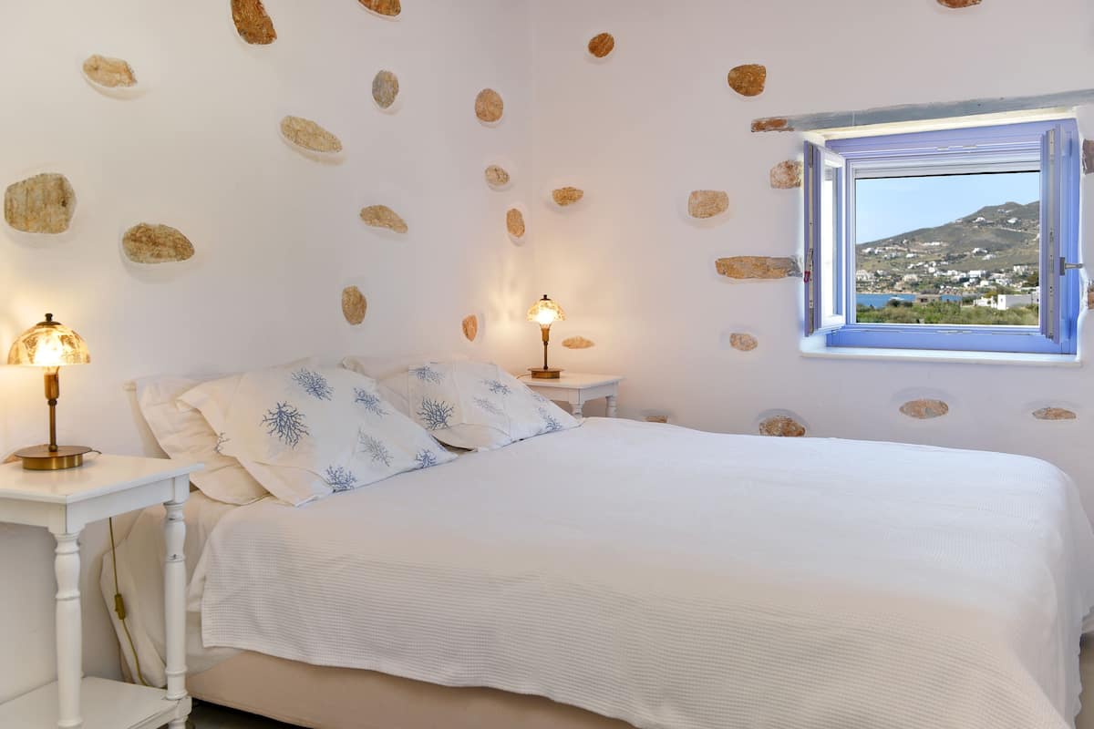 A bedroom with a bed, two bedside tables with lamps, a window with a view of a landscape, and a wall decorated with stones.