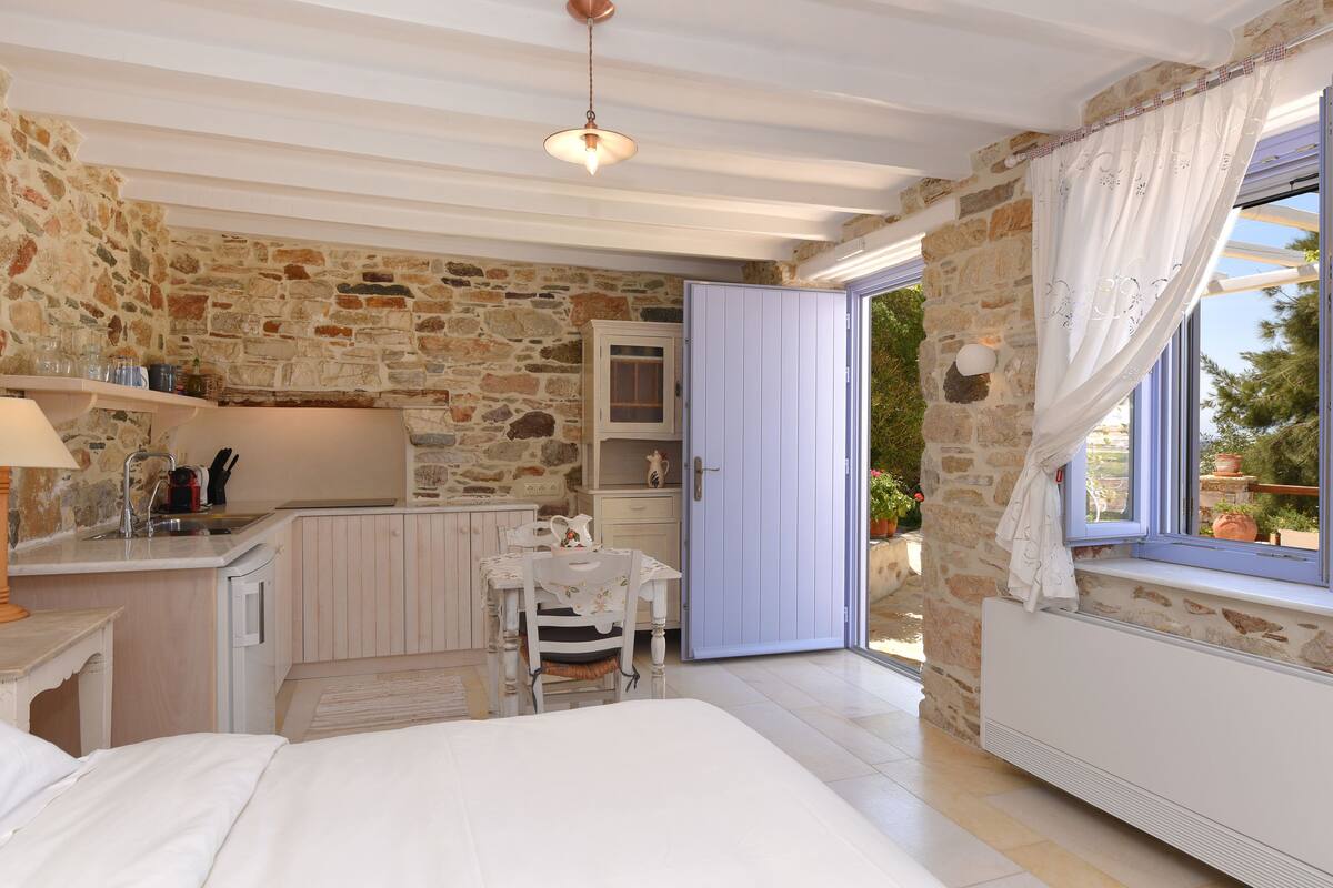 A bedroom with a stone wall, a bed, a small table with a chair, a window with curtains, and a view of the outdoors.