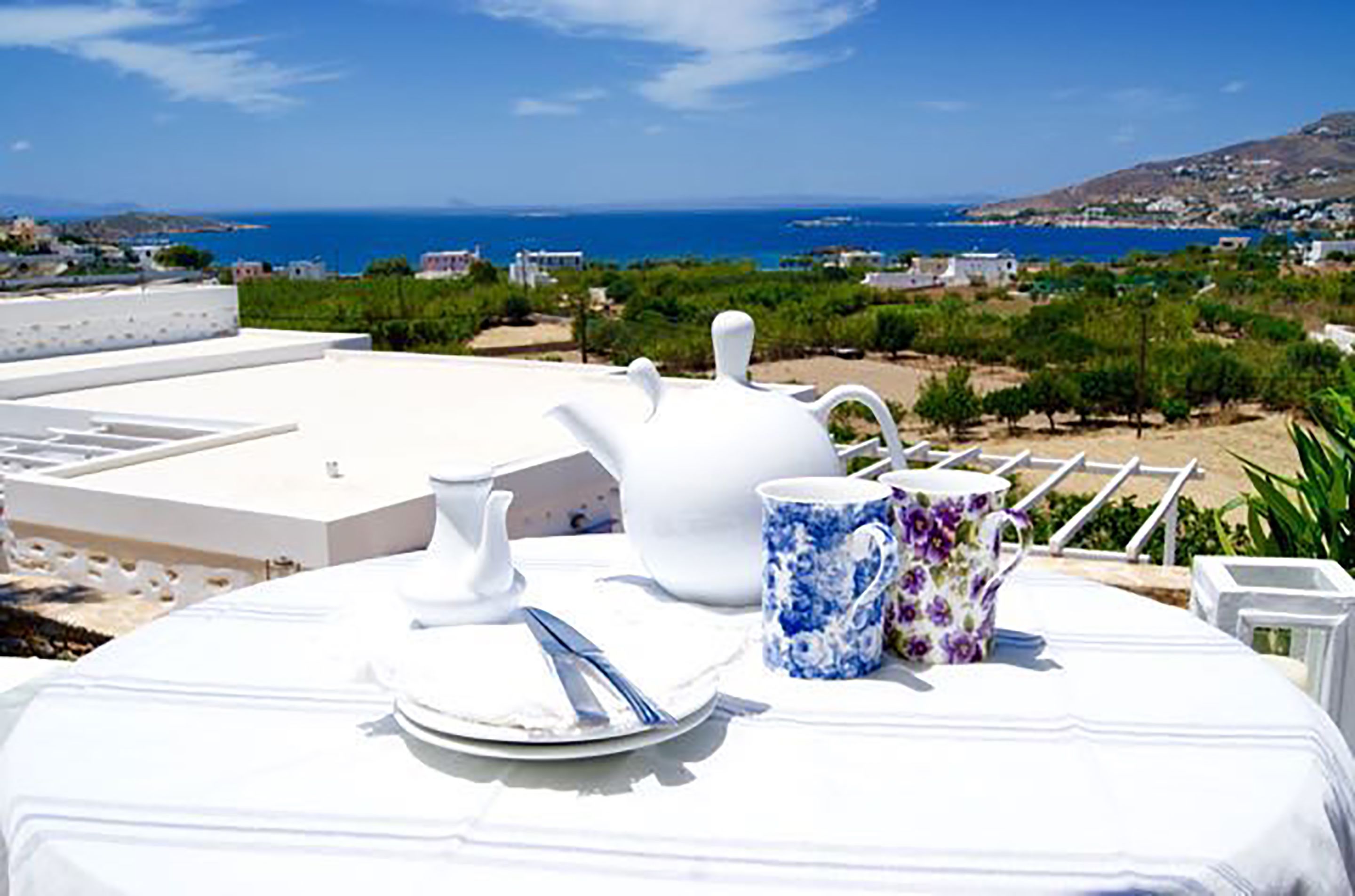 A table set for breakfast with white tableware and a view of the sea and hills.