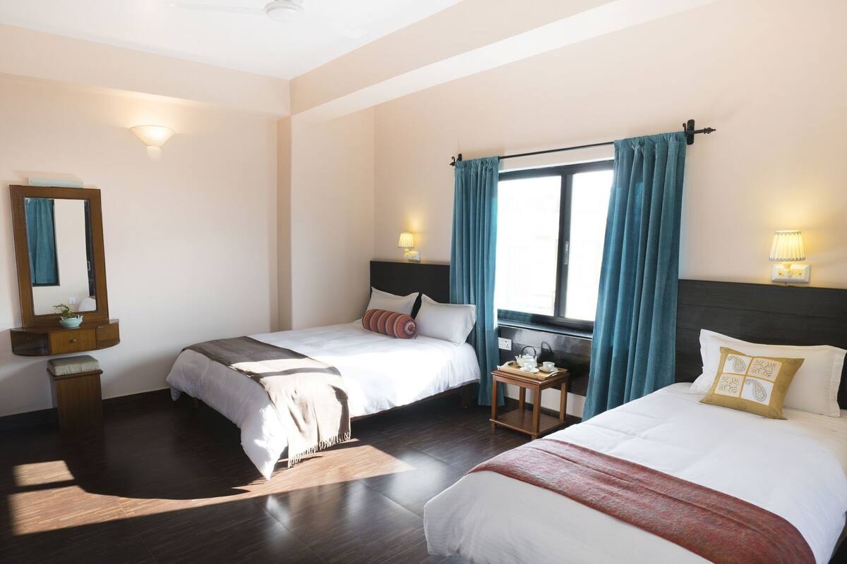 Quarto de hotel com duas camas, uma mesa de cabeceira com abajur, um espelho e uma janela com cortinas.