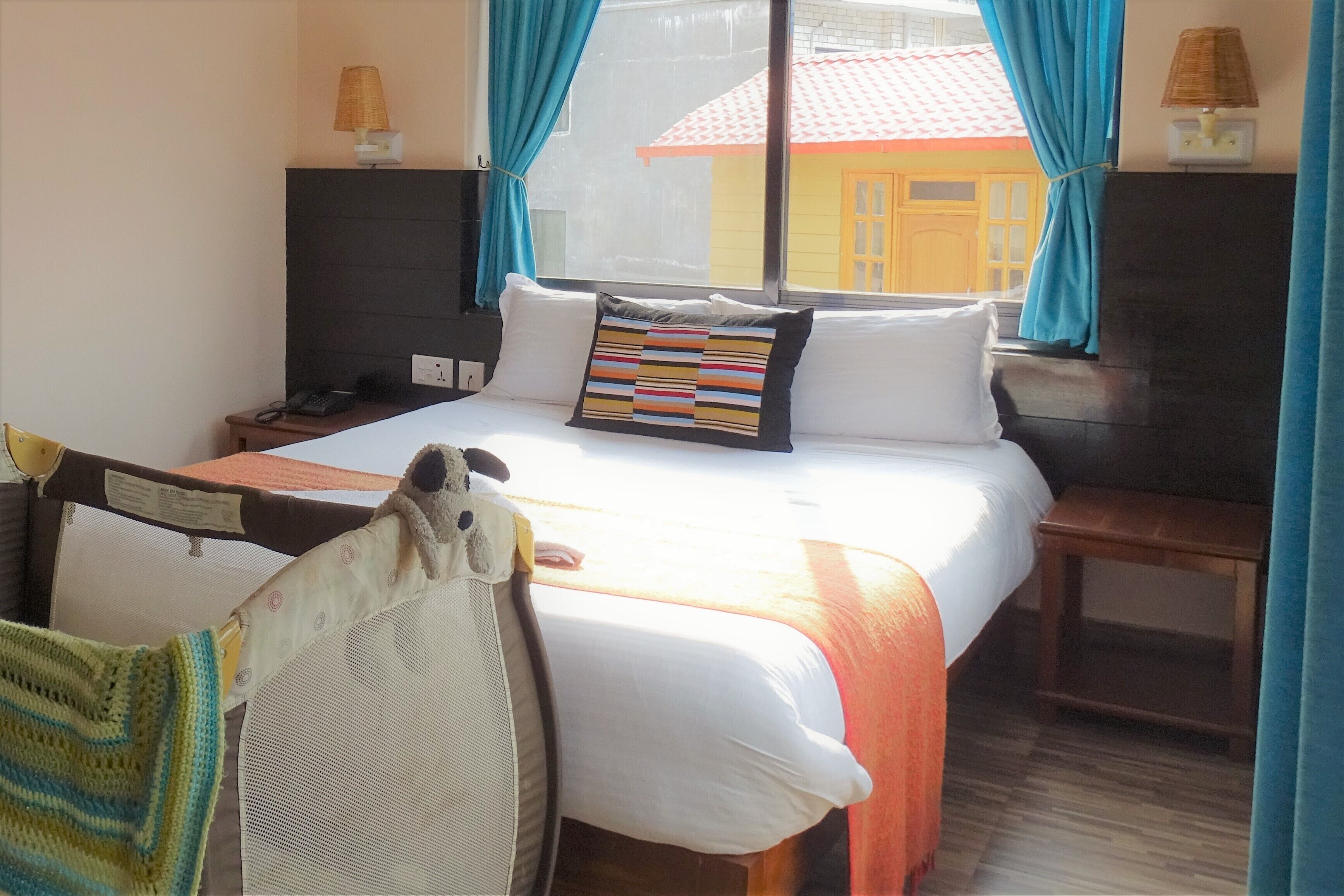 A hotel room with a bed, a nightstand, a lamp, and a view of a building outside.