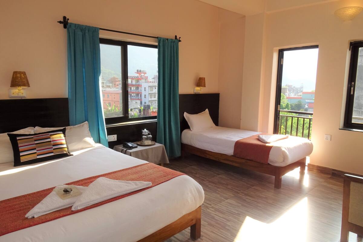 Quarto de hotel com duas camas, uma mesinha com abajur, e vista para edifícios pelas janelas.