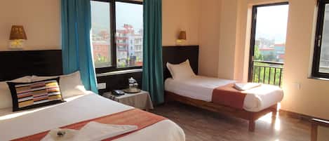 A hotel room with two beds, a small table with a lamp, and a view of buildings through the windows.