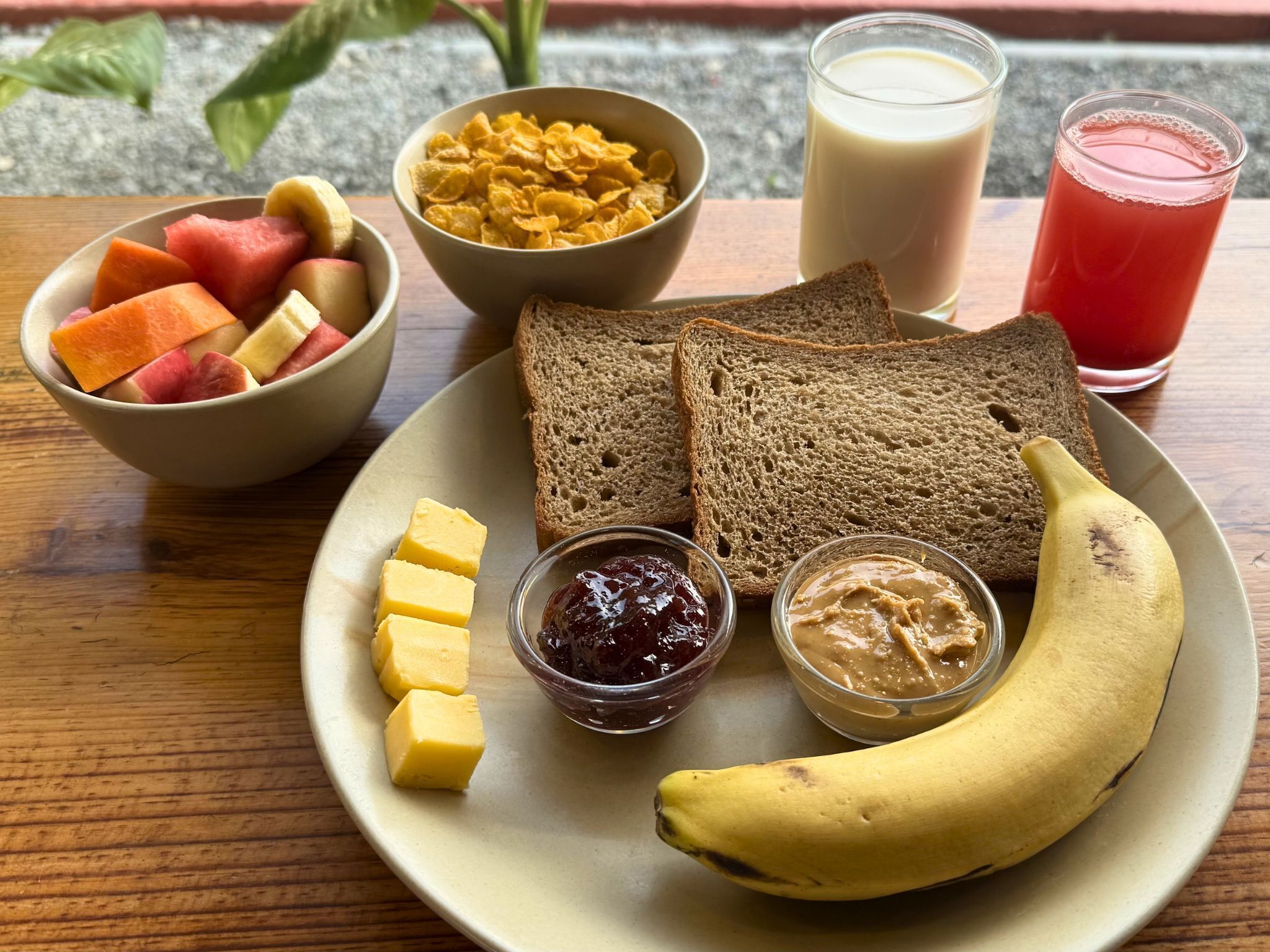 Un plato con banana, pan, mantequilla, mermelada, crema de cacahuate, cereal, leche y fruta.