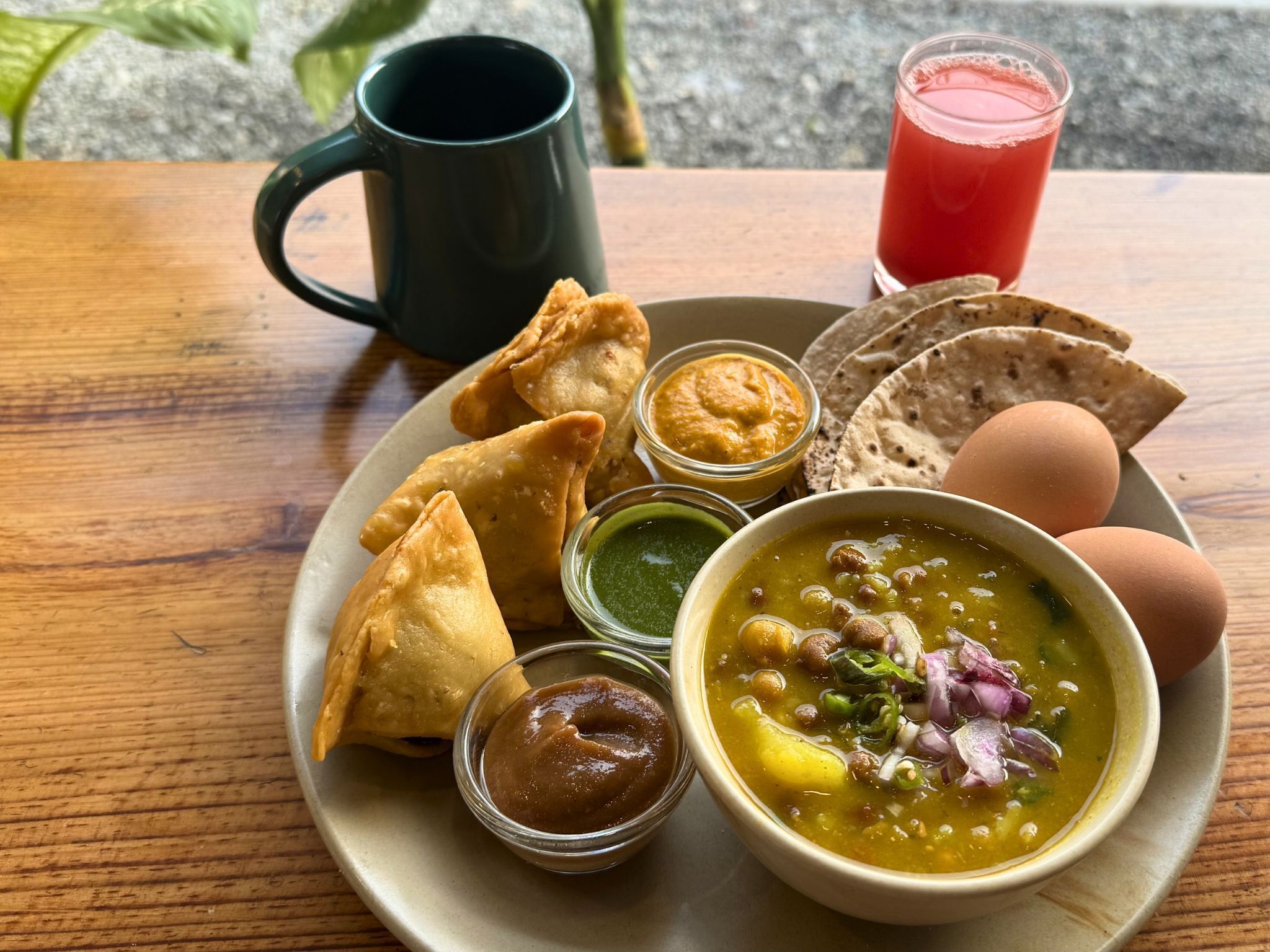 A plate of samosas, chutneys, a bowl of soup, eggs, and a glass of juice on a wooden table.