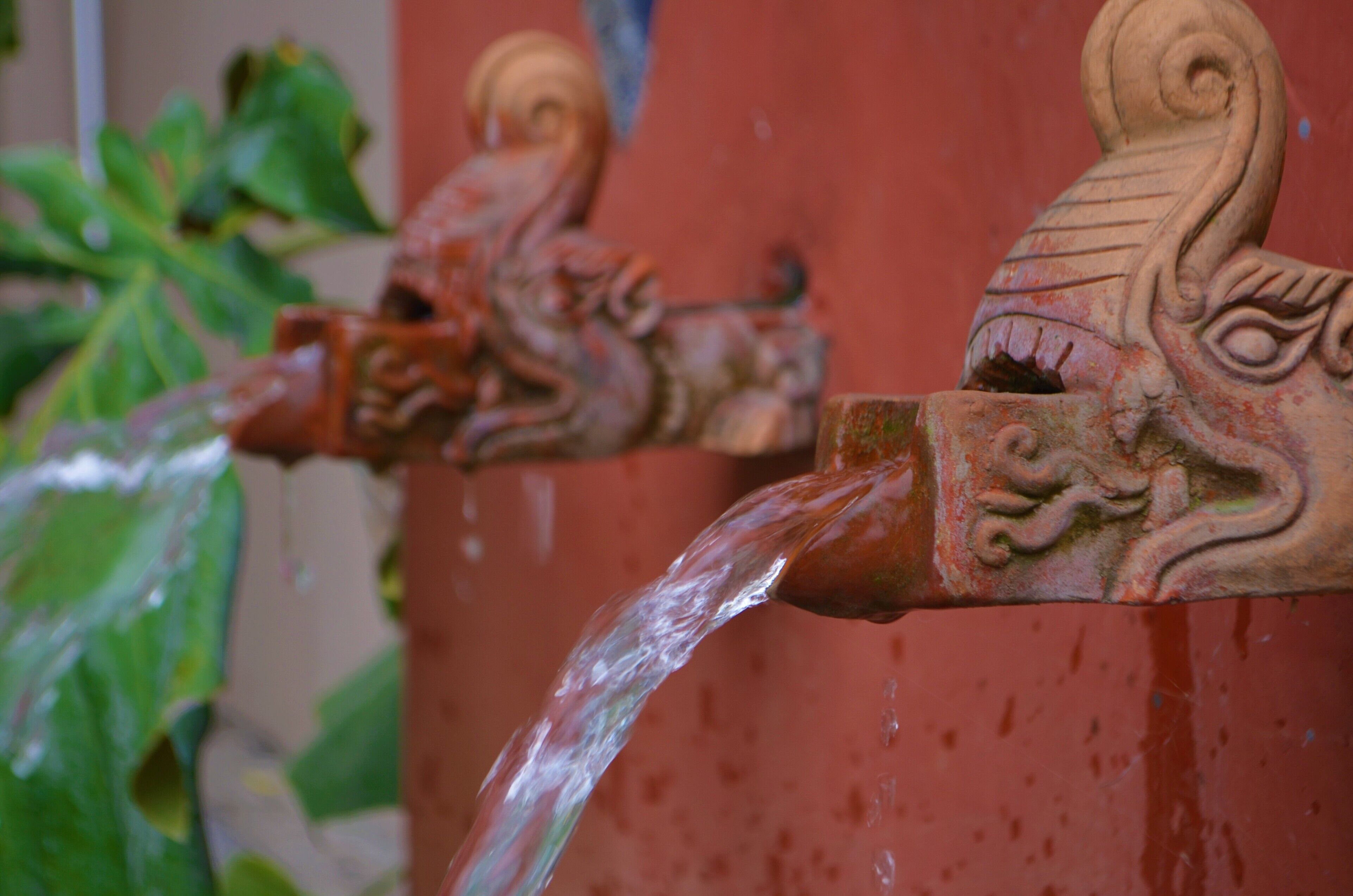 A close-up of ornate, weathered metal water spouts with flowing water, set against a red background.
