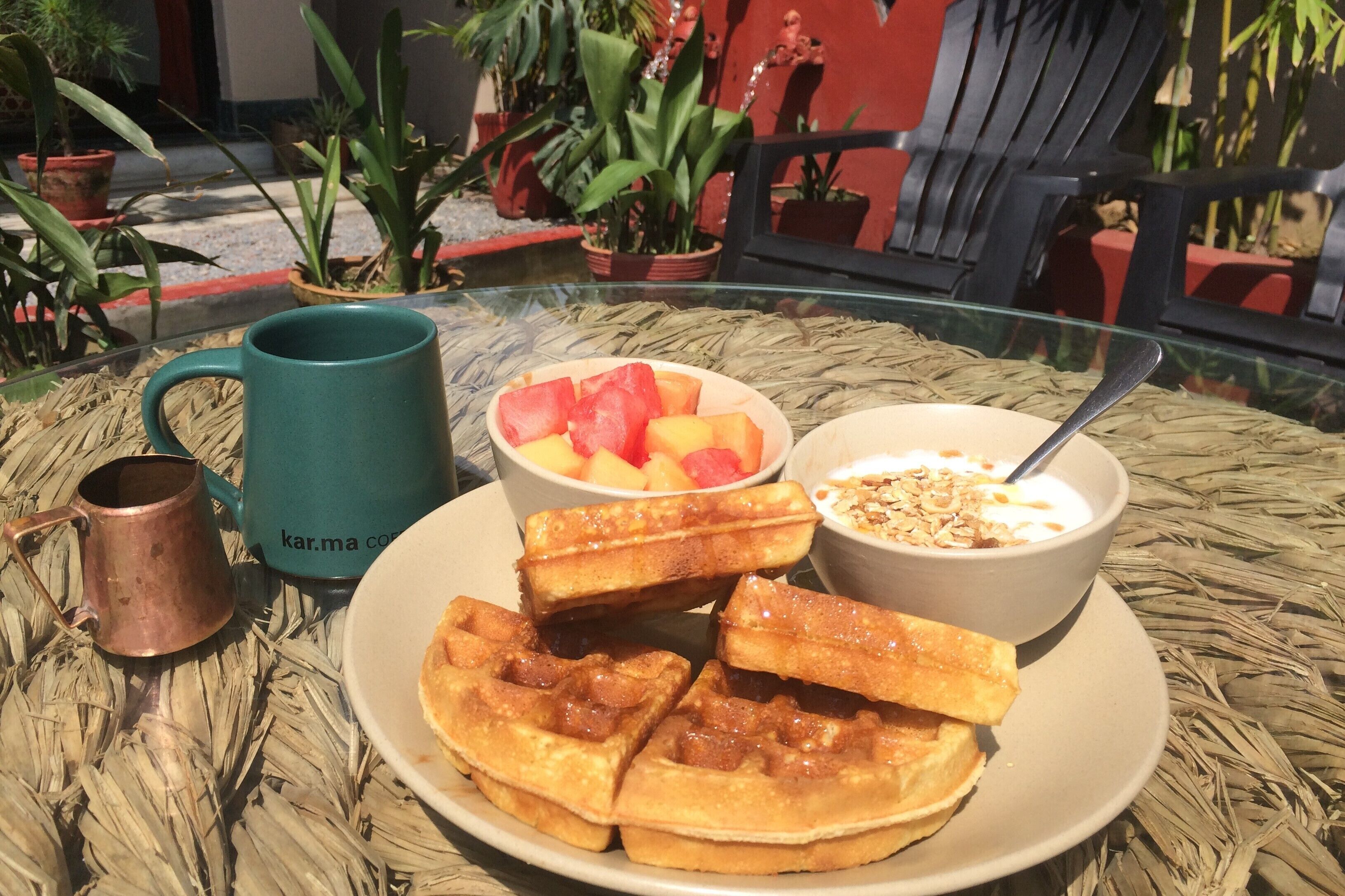 A breakfast setting with waffles, fruit, yogurt, and a mug on a glass table outdoors.
