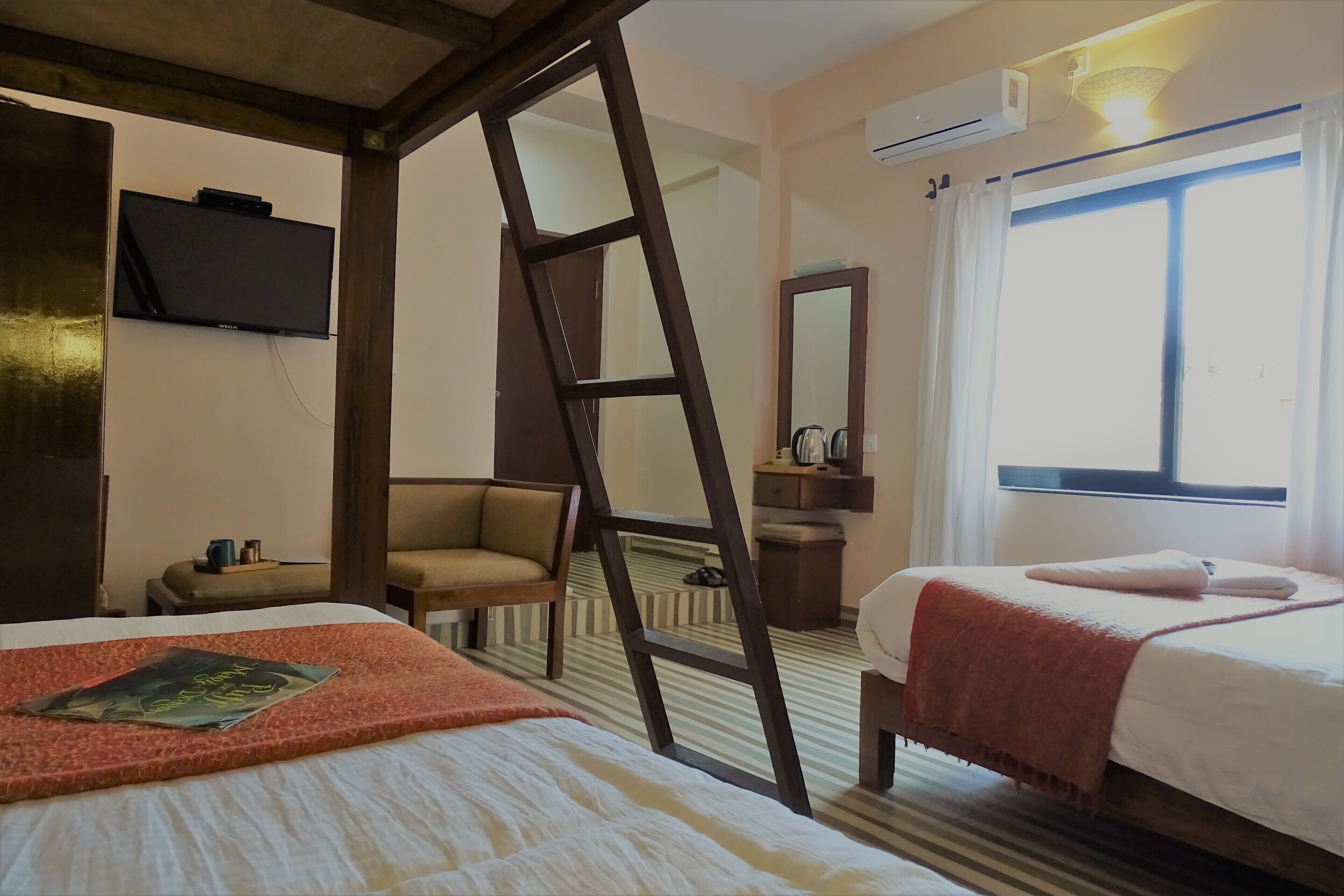 A hotel room with two beds, a TV mounted on the wall, a ladder, a chair, a bedside table, and a window with curtains.