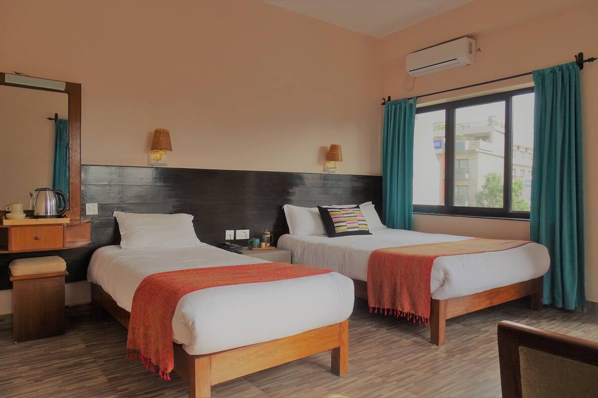 Quarto de hotel com duas camas, cabeceira de madeira, janela com cortinas, mesa de cabeceira com abajur e um aparelho de ar condicionado.