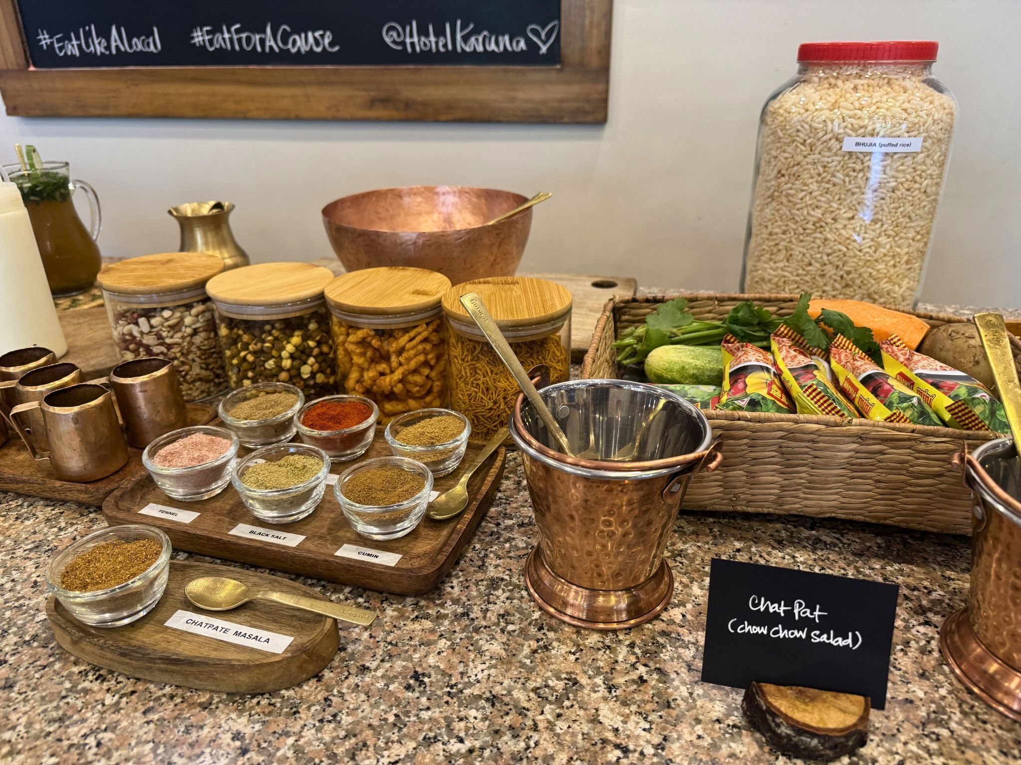 Diversos temperos e ingredientes em uma bancada de cozinha, incluindo recipientes com etiquetas de especiarias como sal negro e cominho, um pote de arroz torrado e vegetais frescos.