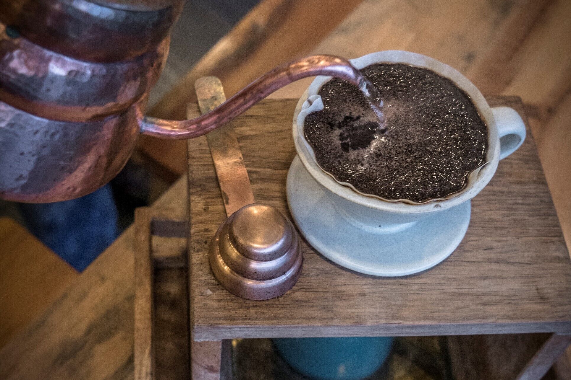 Una tetera de cobre vertiendo líquido en una taza de cerámica blanca sobre una mesa de madera.