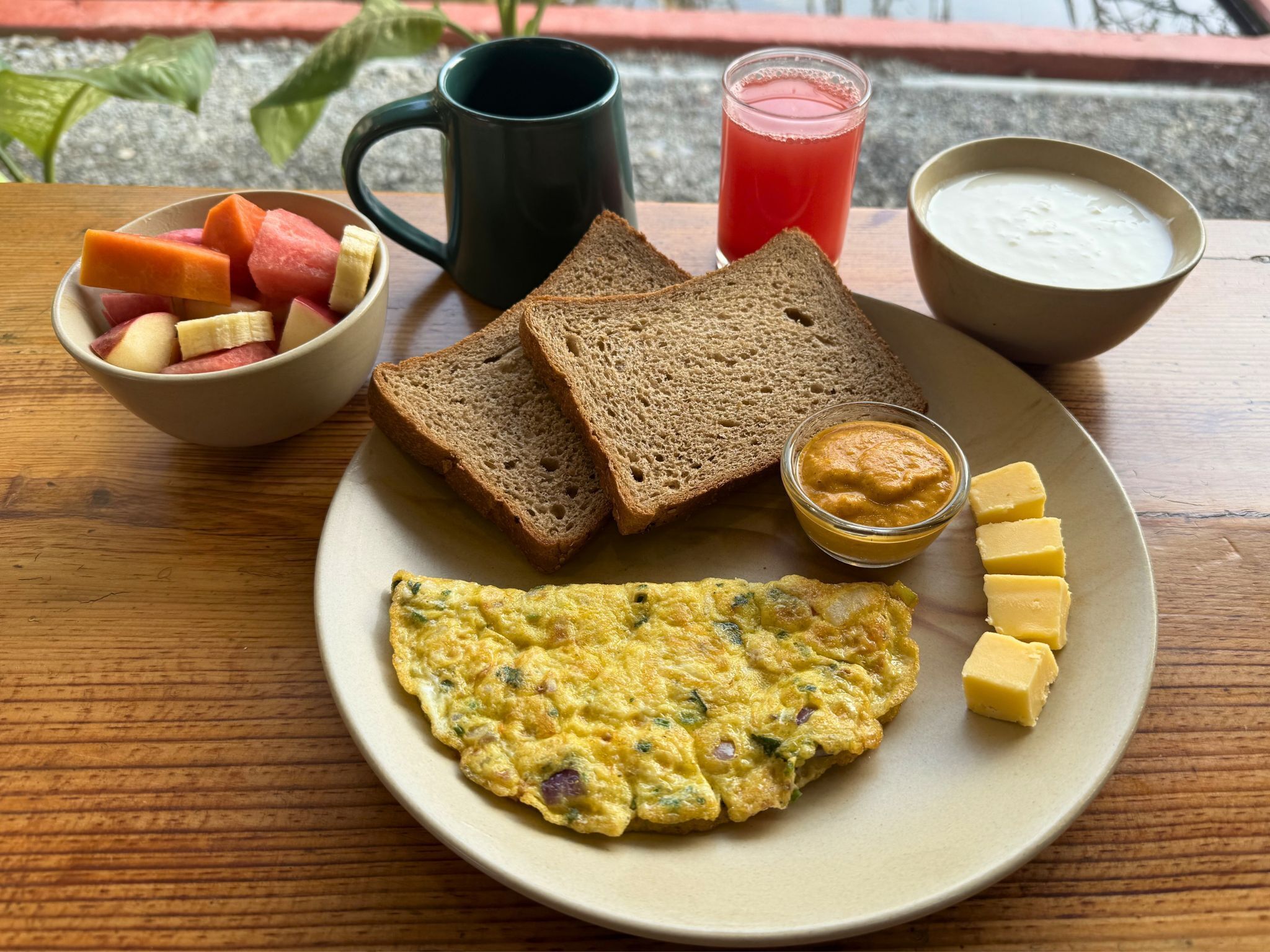 A plate with an omelette, toast, fruit, a mug, a glass of juice, and a bowl of yogurt.