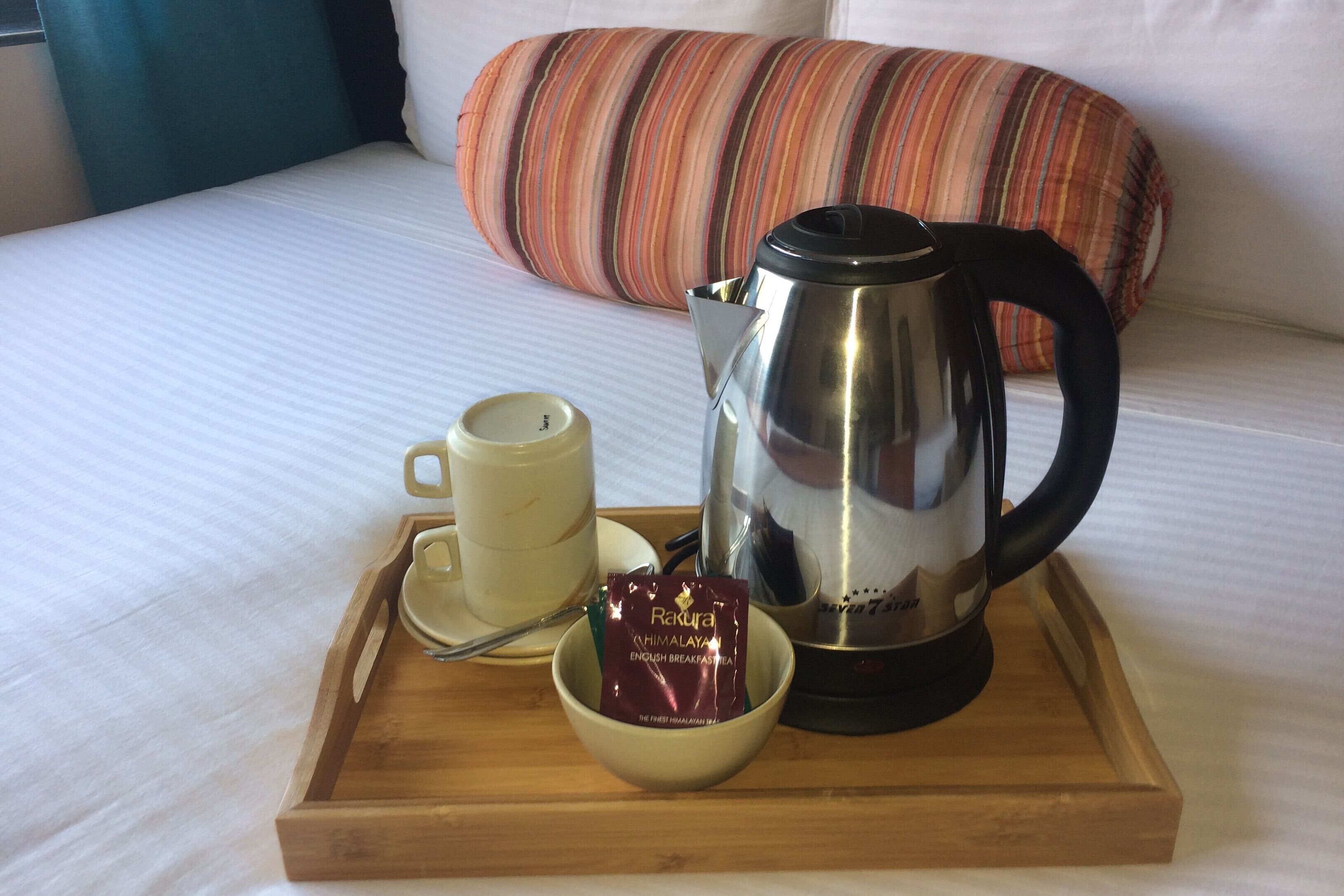 Ein Frühstücksbrett mit Tasse, Schüssel mit Speisen und Wasserkocher auf einem Bett mit gestreiftem Kopfkissen.