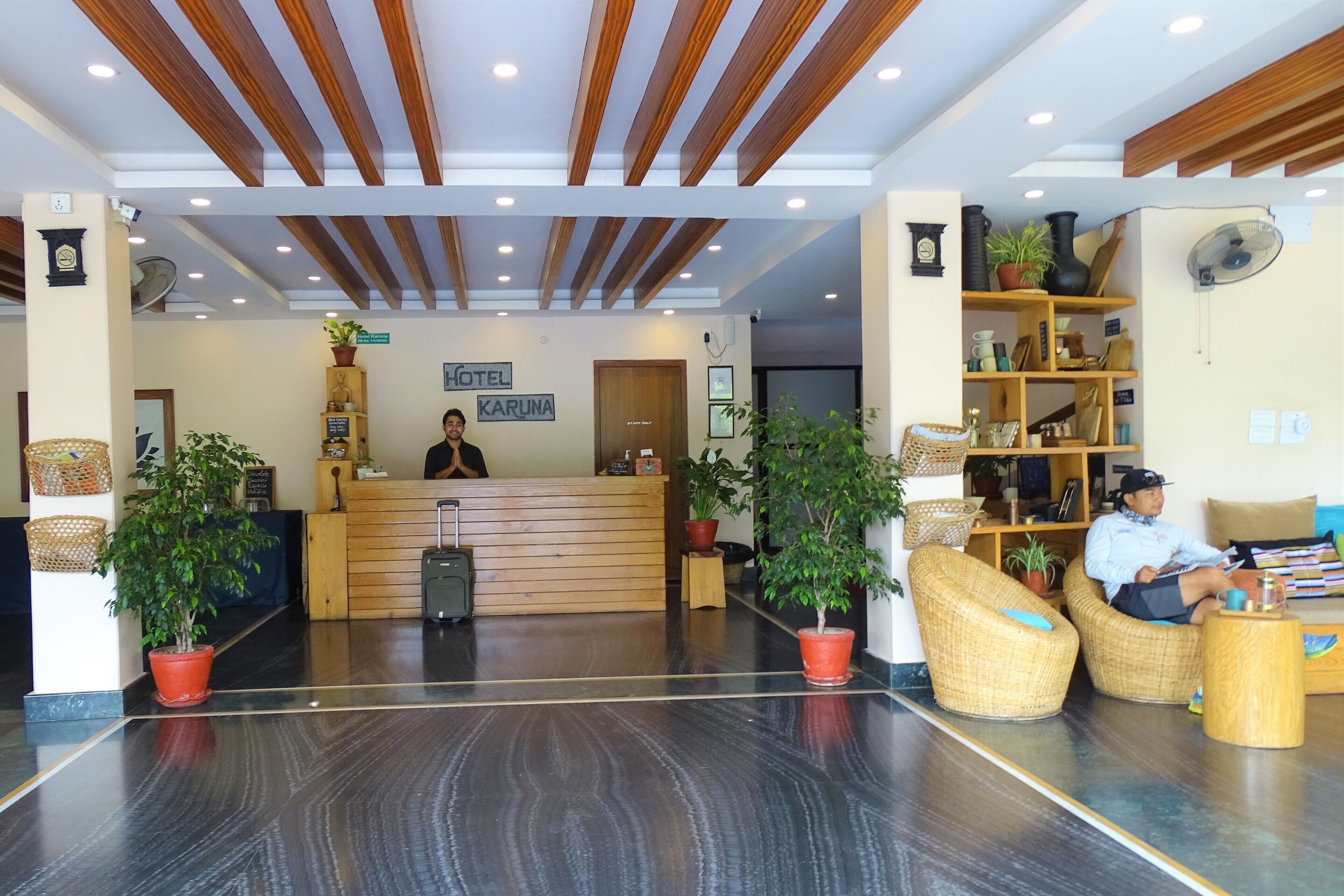 A hotel reception area with a wooden reception desk, a receptionist, wicker furniture, potted plants, and a sign that reads "HOTEL KARUNA".
