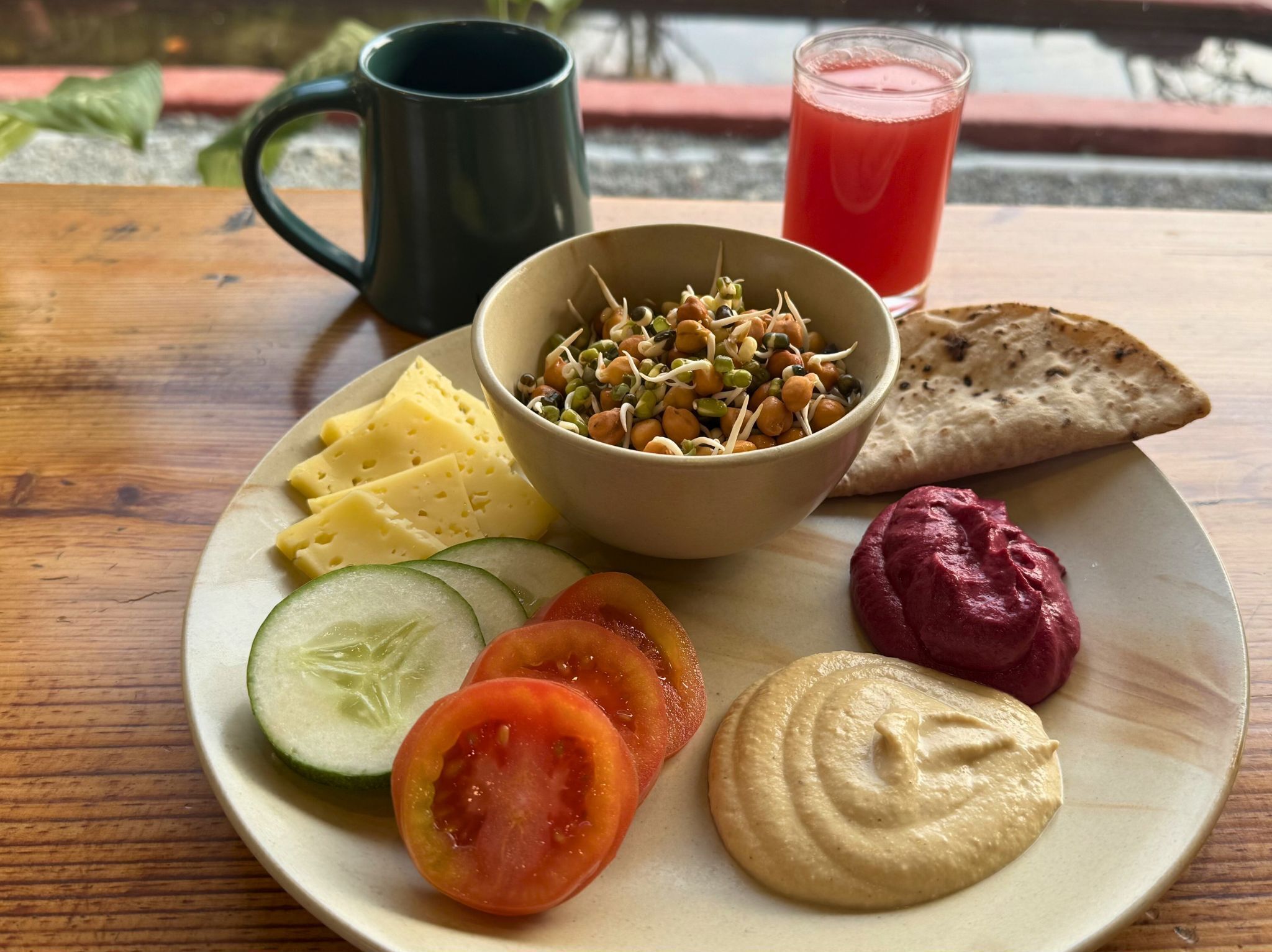 Un plato con queso, brotes, tomate, pepino, hummus y pan plano sobre una mesa de madera.