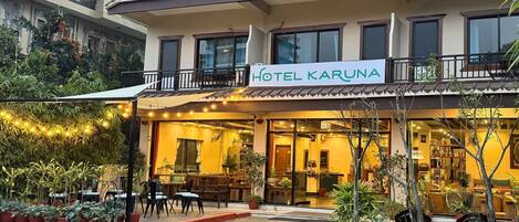 Un hotel con un letrero que dice "HOTEL KARUNA". Cuenta con un balcón con barandilla, plantas en macetas y una zona de estar con sillas y mesas.