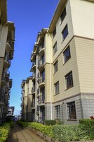 A row of multi-story residential buildings with balconies, a clear sky, and a pathway lined with greenery.