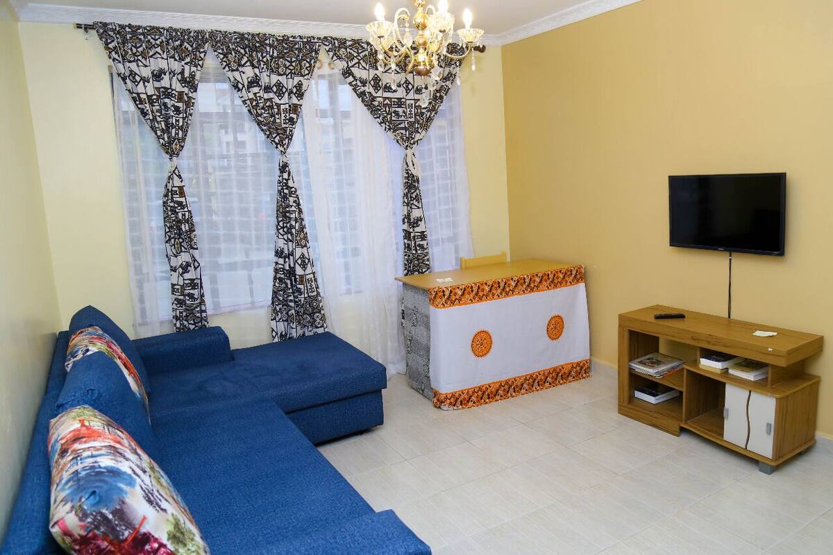 Ein Wohnzimmer mit einem blauen Sofa, einem weißen und orangefarbenen Schrank, einem Fernseher und gemusterten Vorhängen.