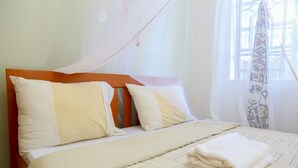 Ein ordentlich bezogenes Bett mit einem gestreiften Bettüberwurf und zwei Kissen, ein Fenster mit einem gemusterten Vorhang und ein leichtes Moskitonetz über dem Bett.