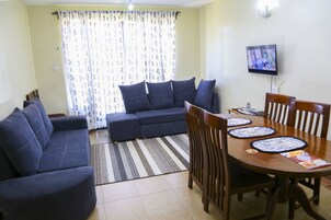 A living room with a sofa, two armchairs, a dining table with chairs, a TV mounted on the wall, and a patterned curtain.