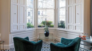 A corner room with a bay window, a glass table, two green chairs, and a vase with plants. - The Lane Hotel (Edinburgh)
