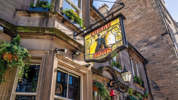 A building with a large, ornate sign above the entrance, a window with a view of the street, and hanging plants. - The Lane Hotel (Edinburgh)