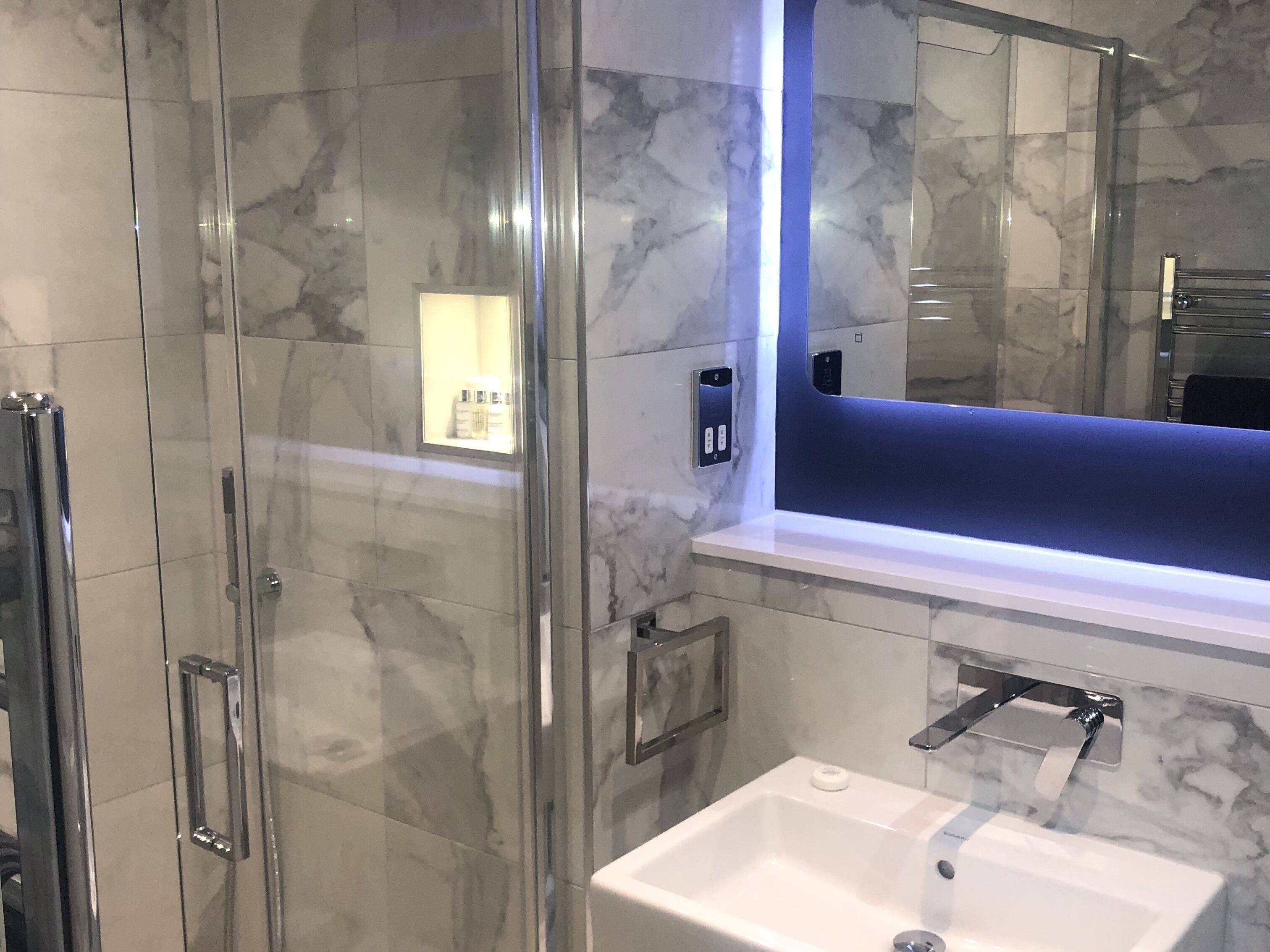 Une salle de bain moderne avec une douche à parois de verre, un lavabo blanc et un espace coiffeuse éclairé en bleu.