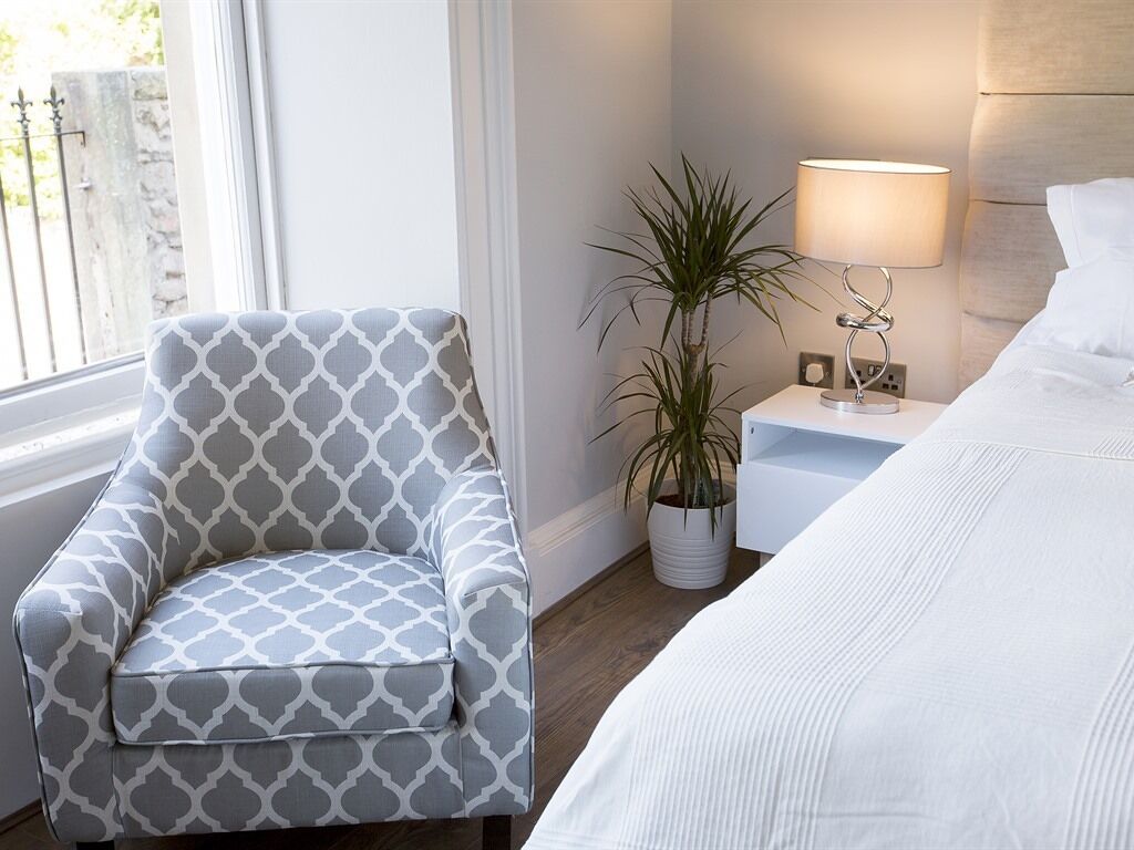 Hotellihuone, jossa on kuviollinen nojatuoli, yöpöytä lampun kanssa, ruukkukasvi ja valkoisilla lakanoiden kanssa varustettu sänky.