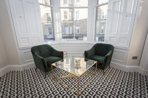 Een kamer met een tafel met glazen blad, twee groene fauteuils, een kamerbreed tapijt en grote ramen met uitzicht op gebouwen.