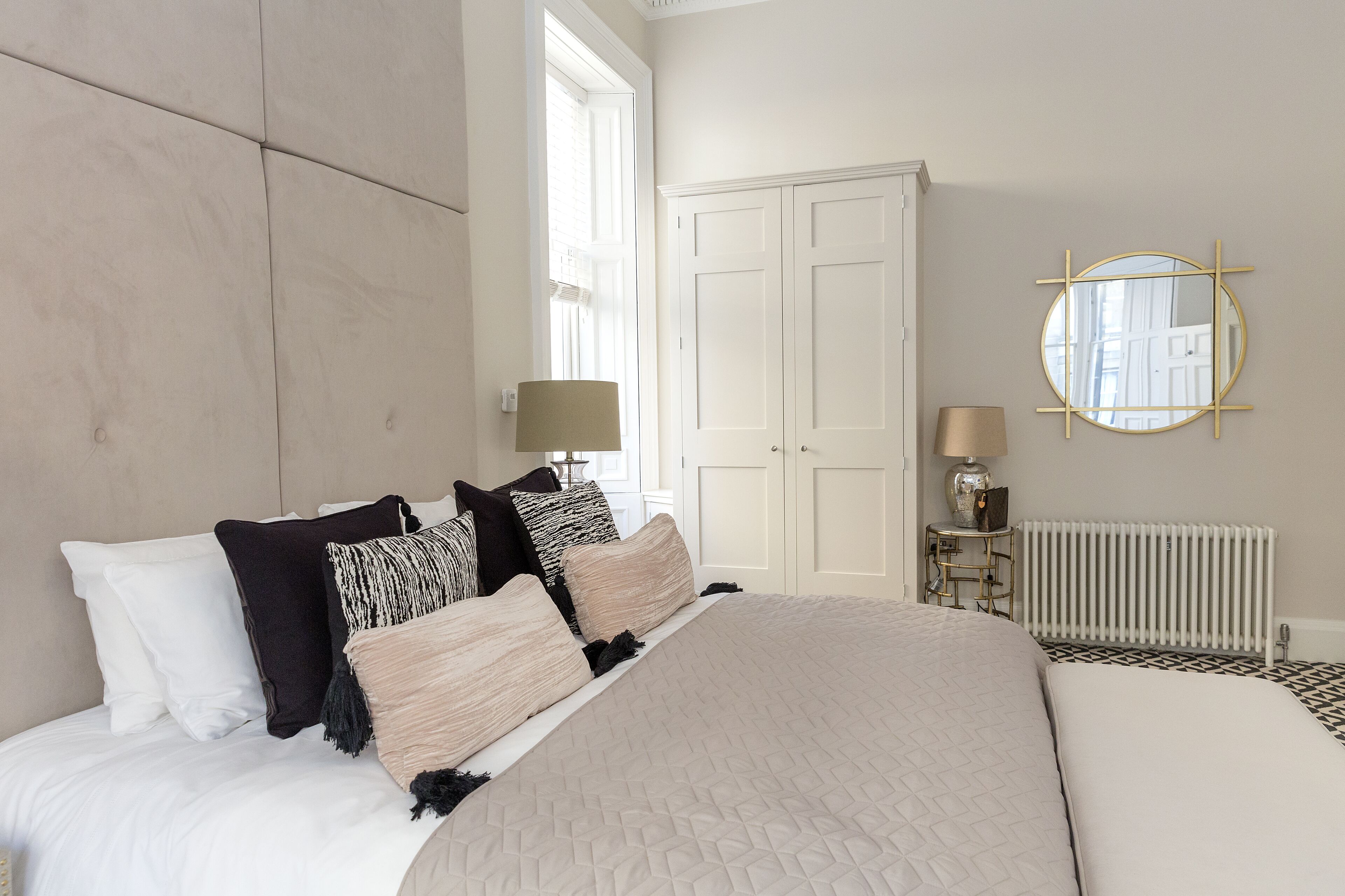 A bedroom with a large bed, white walls, a white wardrobe, a round mirror, and a radiator.