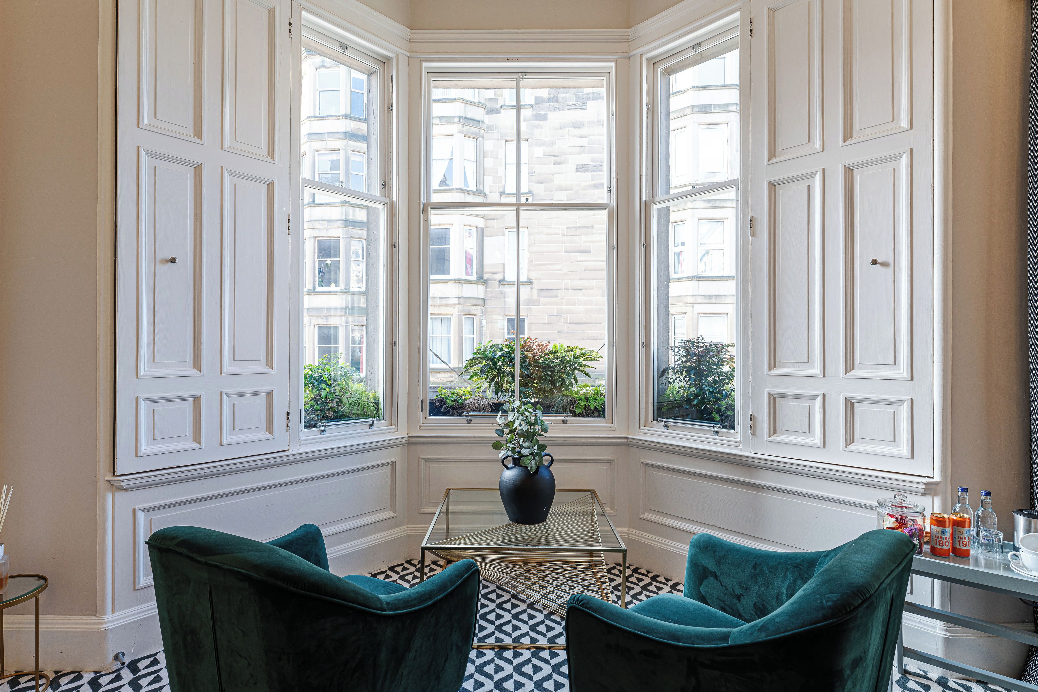 A corner room with a bay window, a glass table, two green chairs, and a vase with plants.