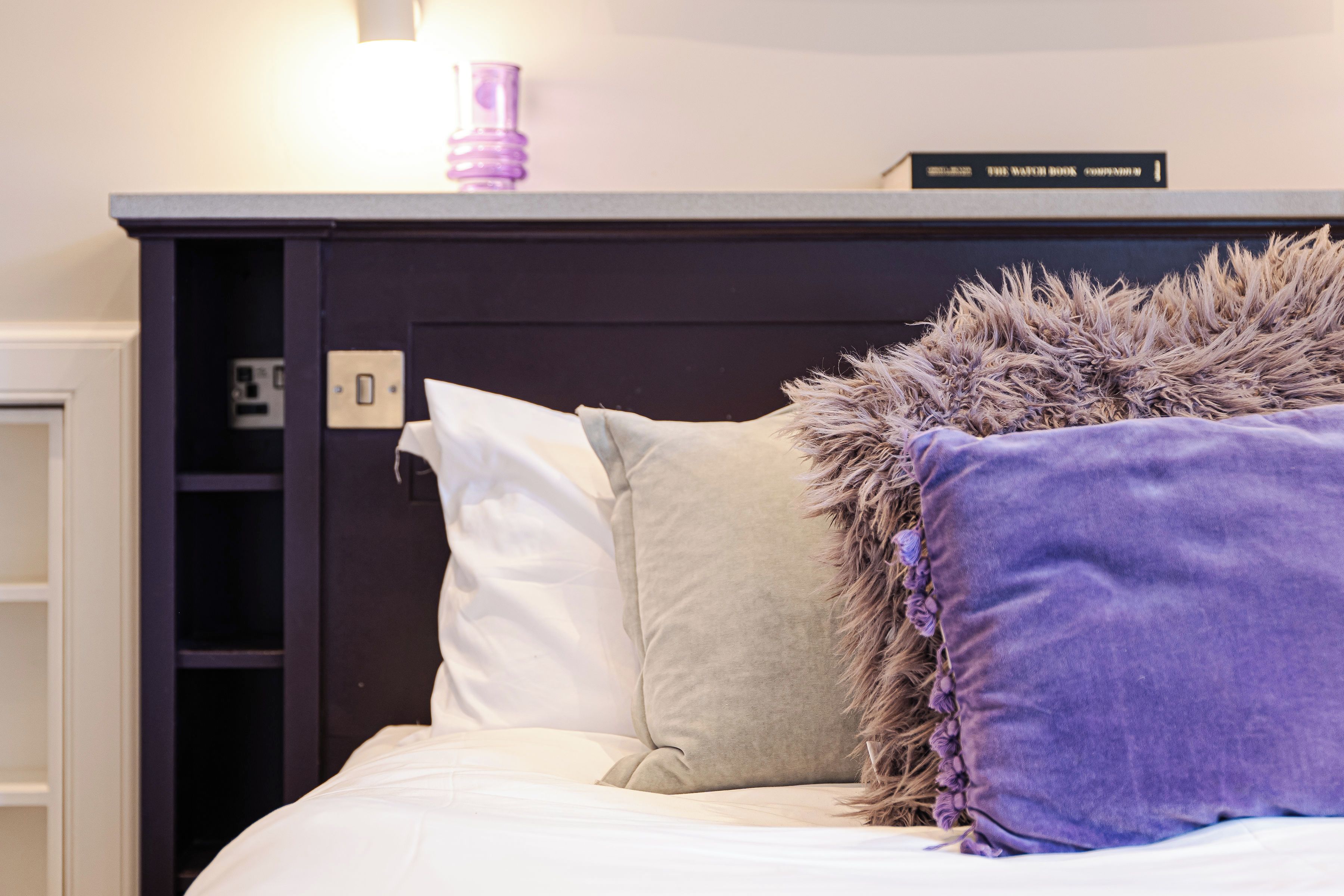A bed with white and purple pillows, a dark headboard, and a small shelf with a book and a lamp.