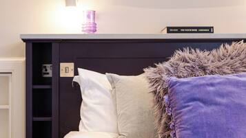 A bed with white and purple pillows, a dark headboard, and a small shelf with a book and a lamp.