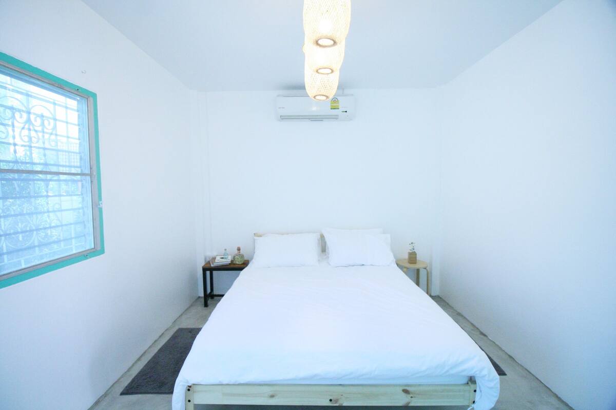 Et lille, rent værelse med en enkeltseng, et natbord, et vindue og en aircondition.