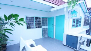 A bright, clean room with white walls, a teal door, and a window with decorative bars. There are white plastic chairs and a potted plant. - Hello Hostel Pakchong (Pak Chong)