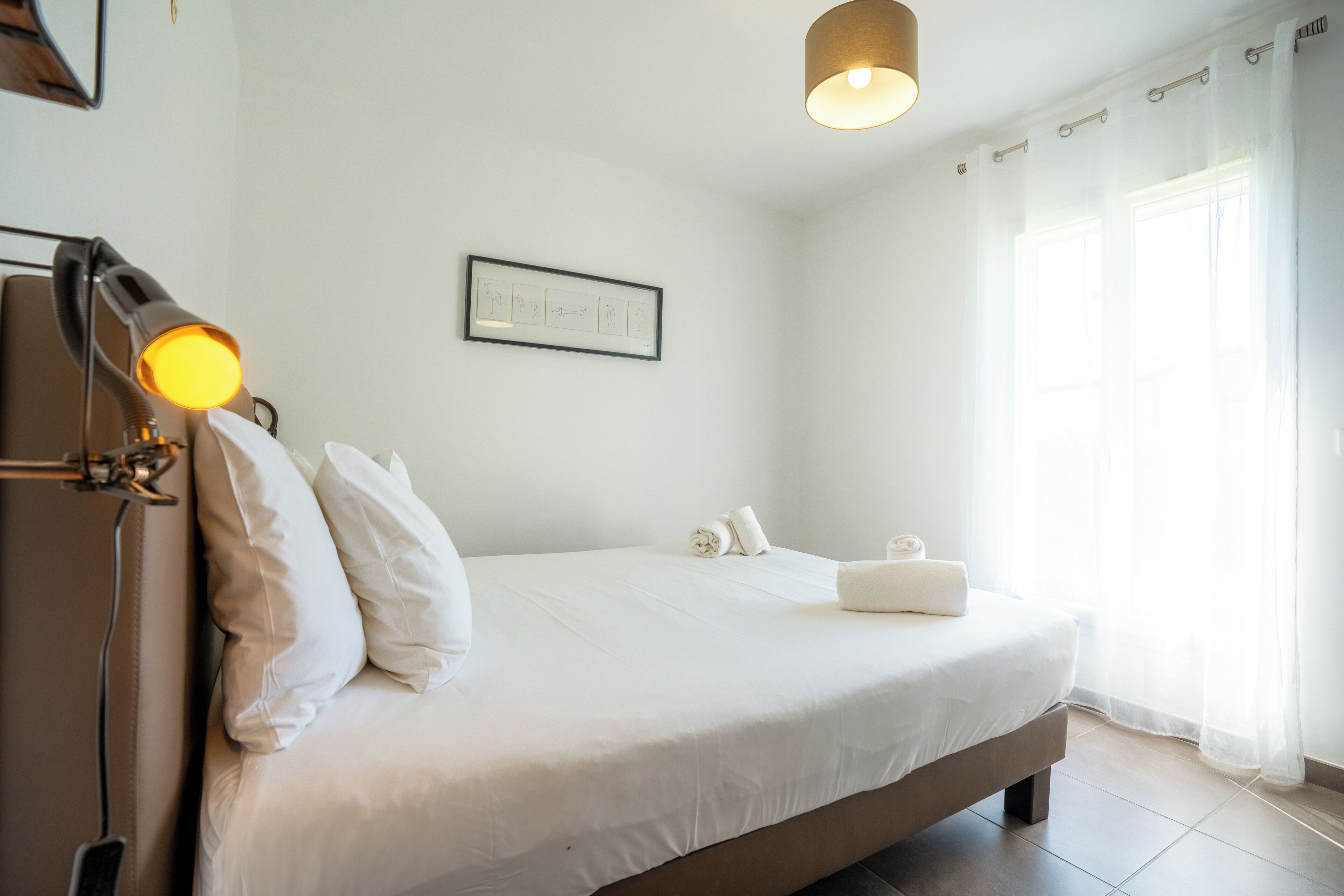 Una cama bien hecha con sábanas blancas, una lámpara de noche y un cuadro en la pared.
