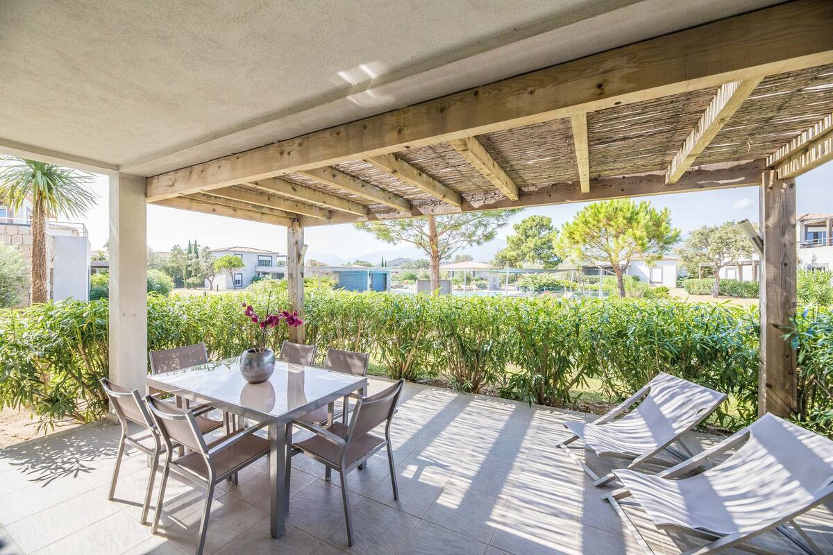 A covered patio with a table and chairs, surrounded by greenery and a view of the neighborhood.