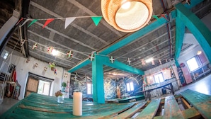 A room with a large, round, woven light fixture, teal structural beams, and a wooden table with a white candle. - Surf Camp San Sebastian Partyhostel (Aia)