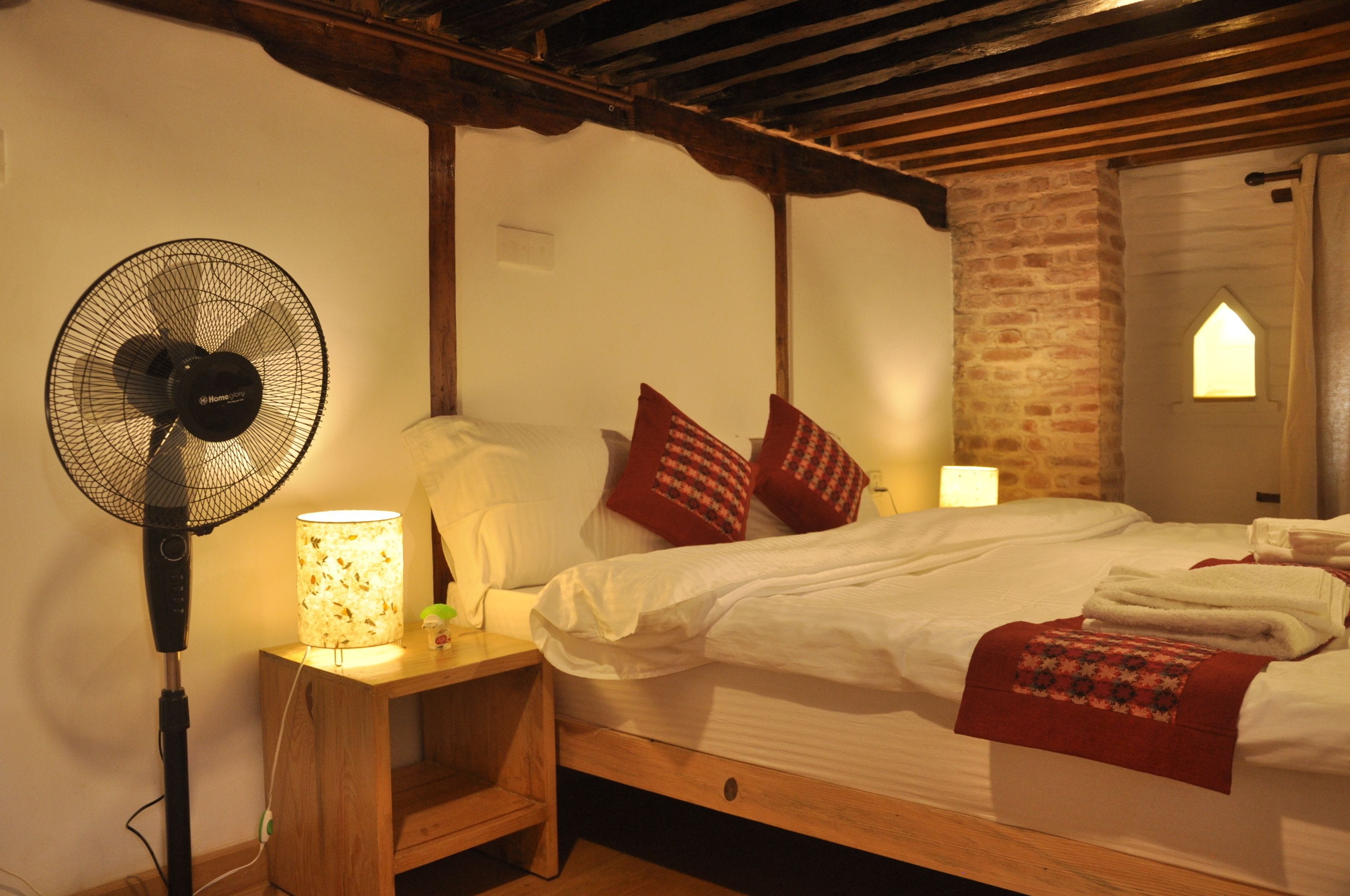 Una camera da letto con un letto, una ventola da pavimento, un comodino e un muro in mattoni.