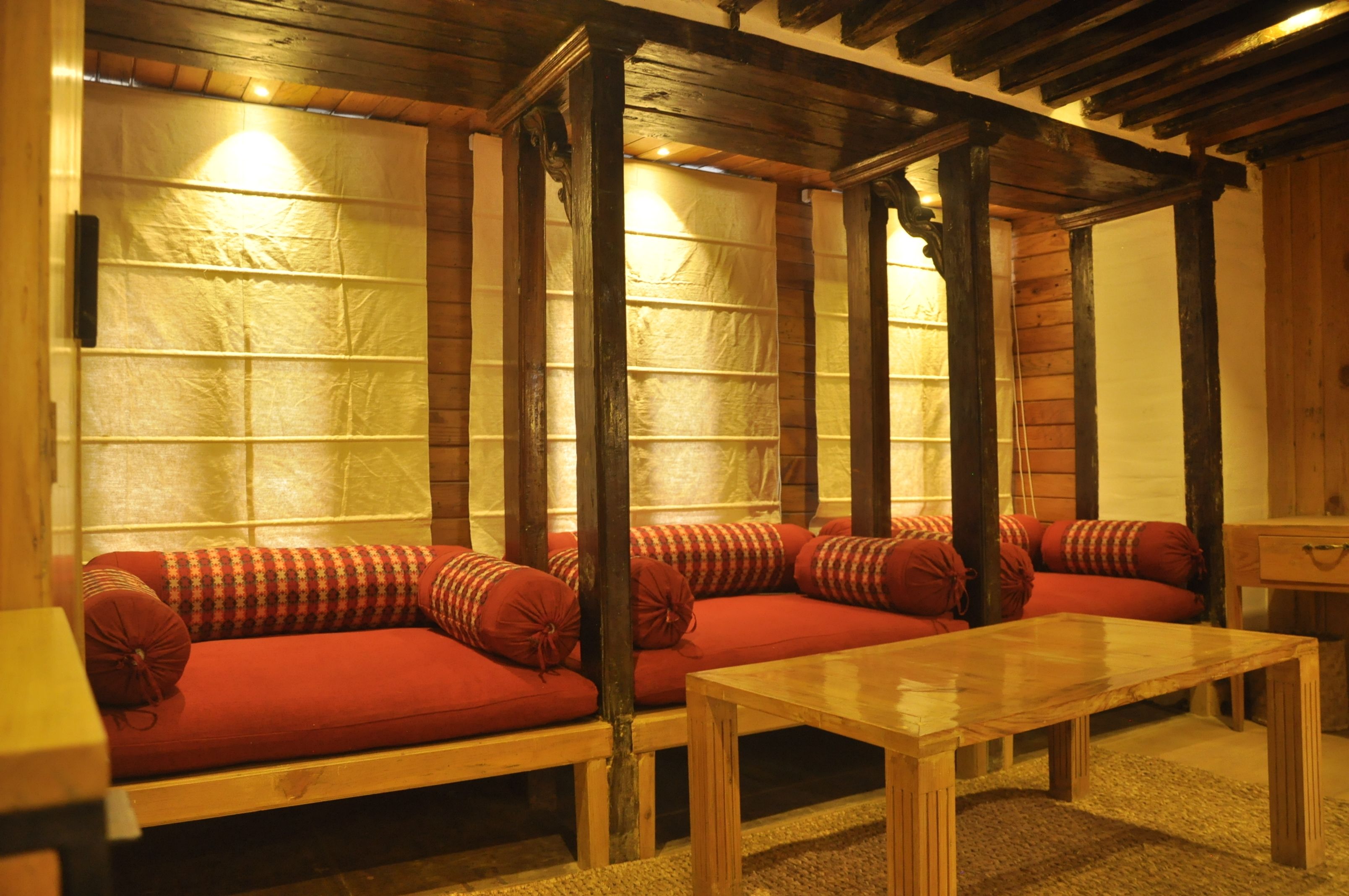 A cozy room with a wooden bench and a table, a red sofa with patterned cushions, and a wooden ceiling structure.