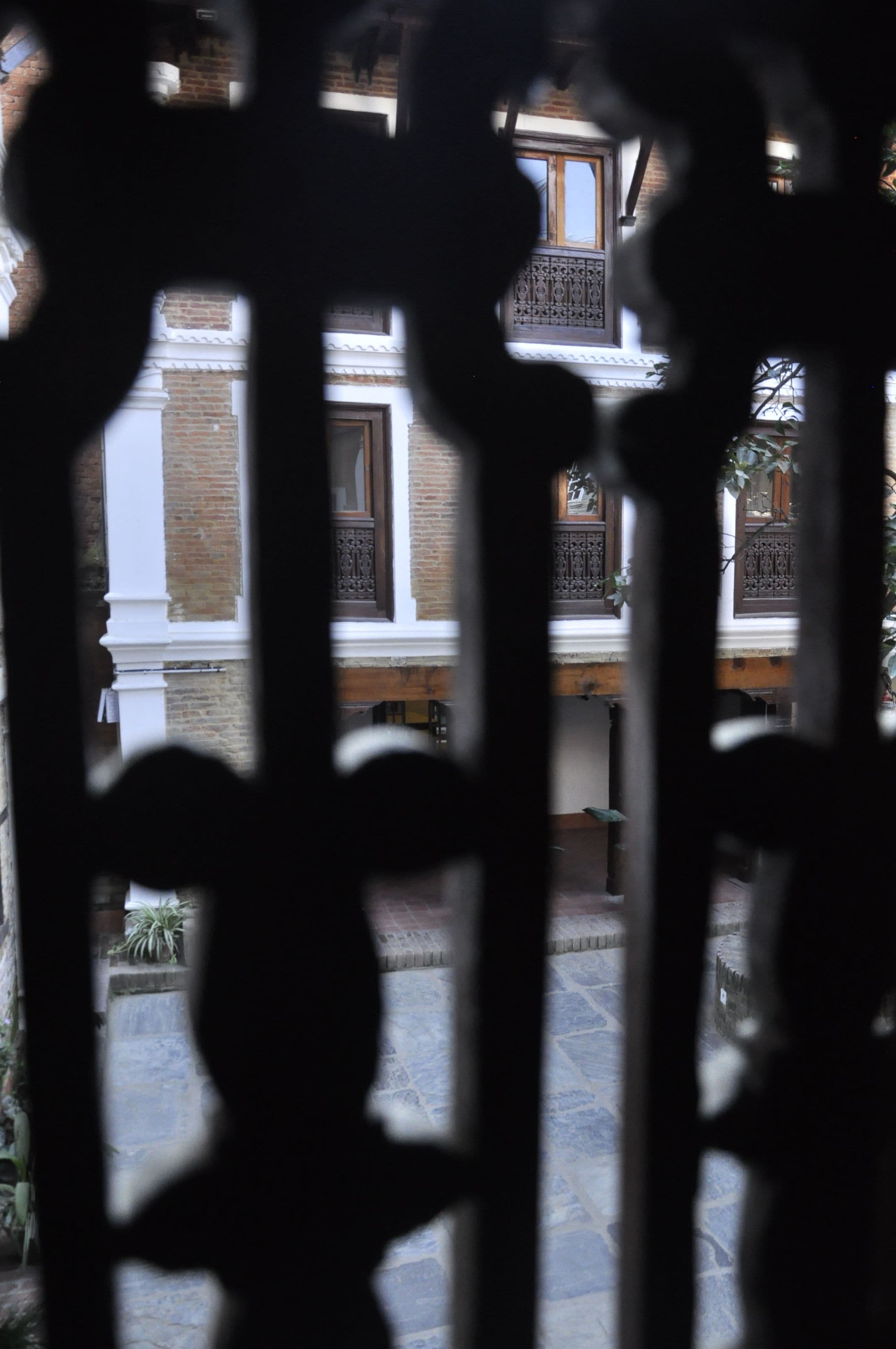 Vista a través de elaboradas rejas de hierro de un edificio con múltiples ventanas y un patio empedrado.