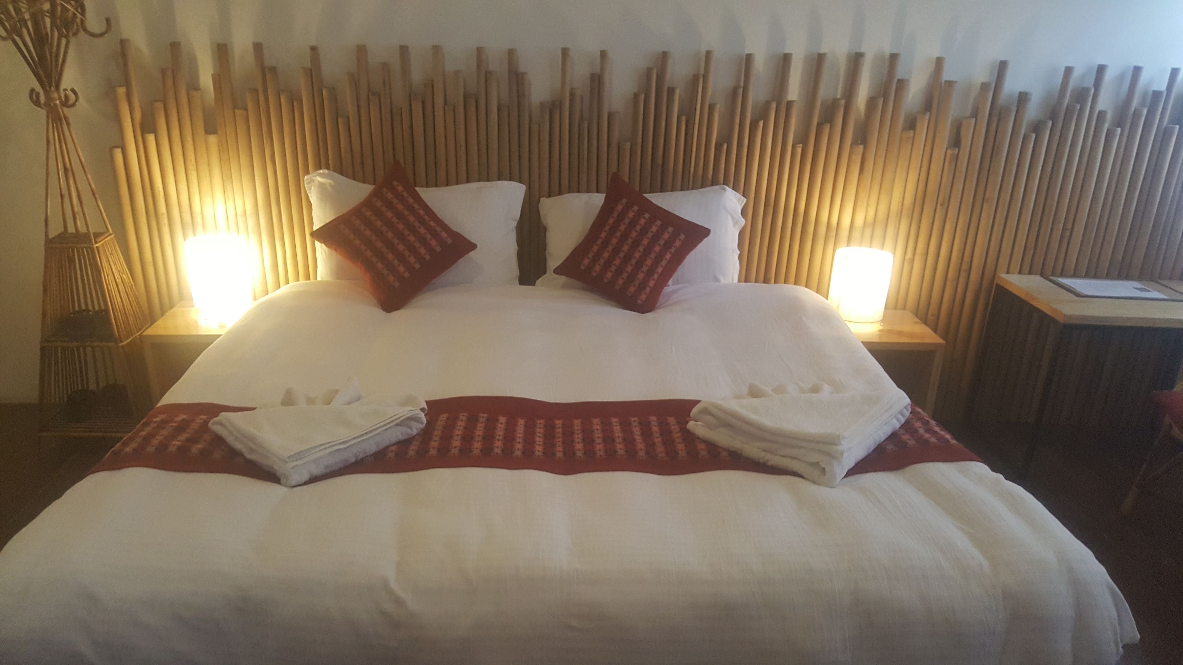 Una cama bien tendida con sábanas blancas y detalles rojos, una mesita de noche con lámpara, y una pared con listones de madera verticales.